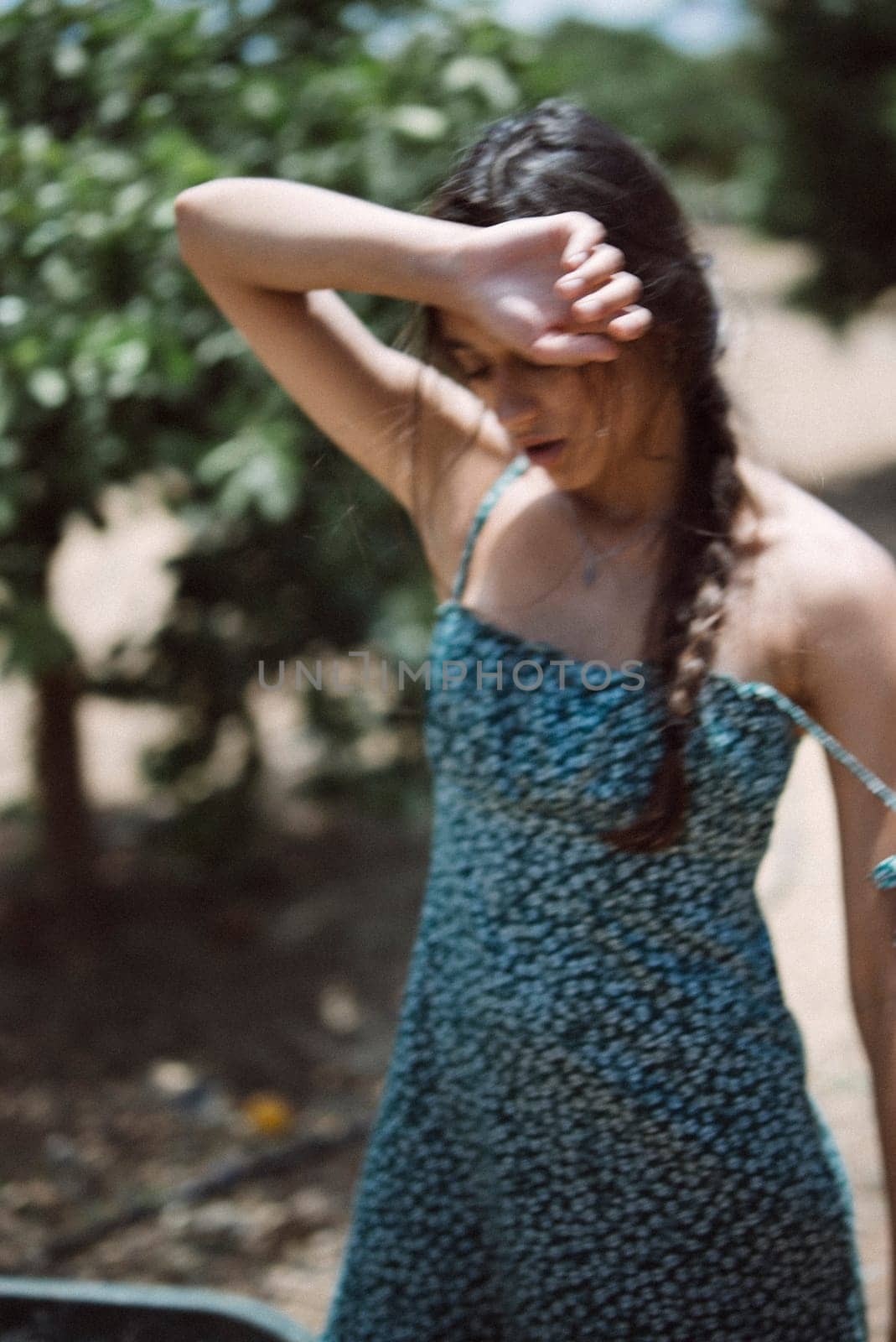 A young woman in a patterned dress stands outside in an orchard, shading her face from the sun with her hand
