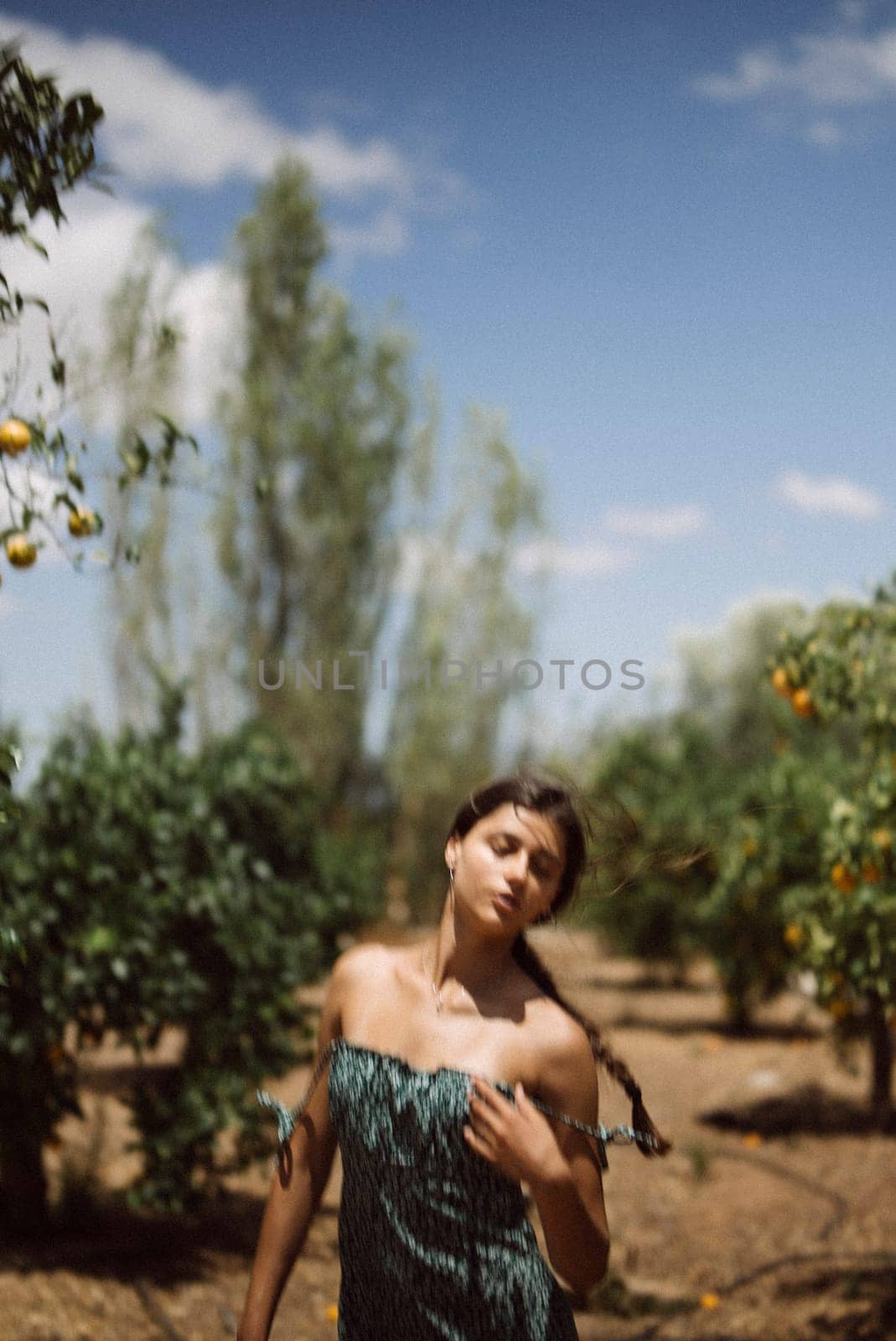 A tranquil scene of a young woman in a green dress standing in an orchard with fruit trees under a clear blue sky