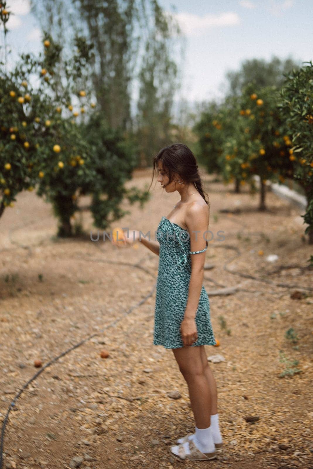 A woman in a green dress and sandals is standing in an orchard, holding an orange while surrounded by trees.