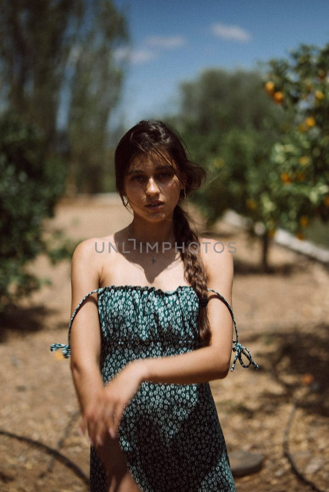 A young woman with braided hair posing in a blue summer dress, surrounded by nature in a sunny outdoor setting