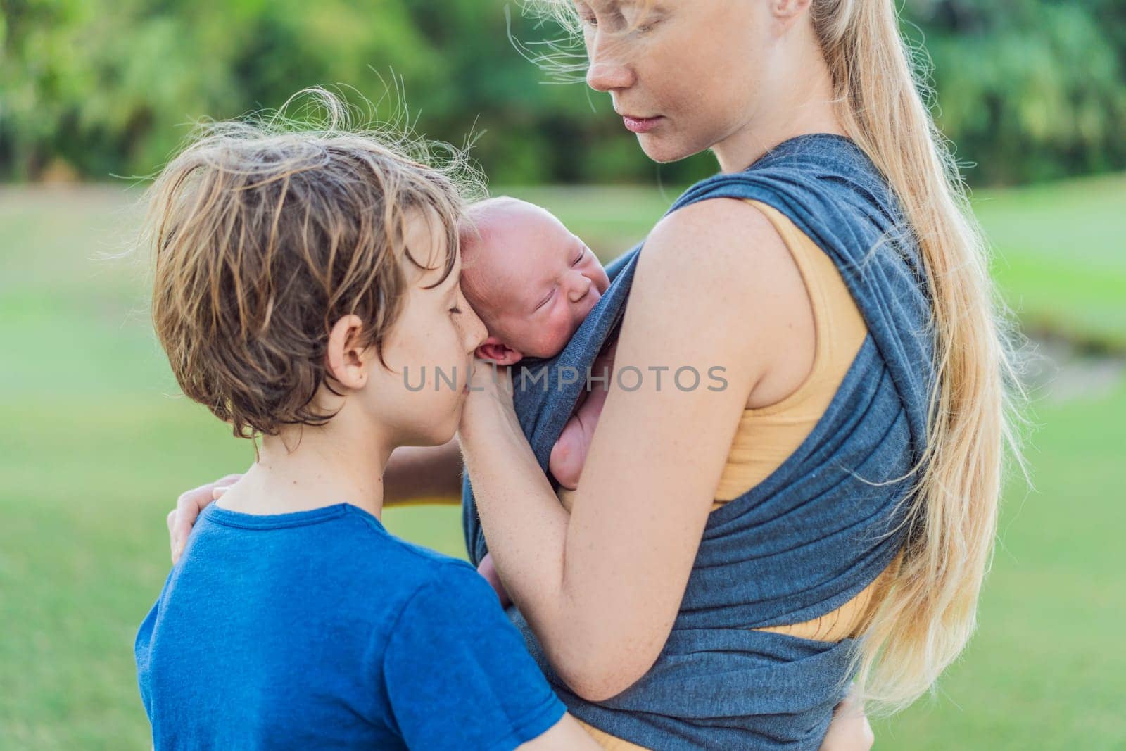 Mom walks with a newborn in a sling and also with her teenage son. This scene showcases the bond within the family, highlighting the nurturing care of a mother and the connection between siblings.