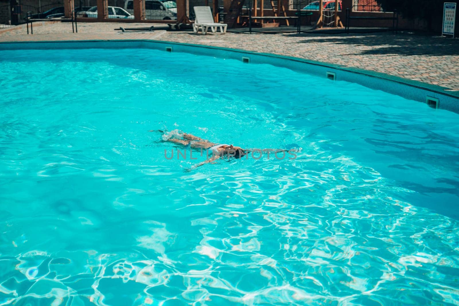 A woman is swimming in a pool. The pool is surrounded by a fence and has a playground in the background. by Matiunina