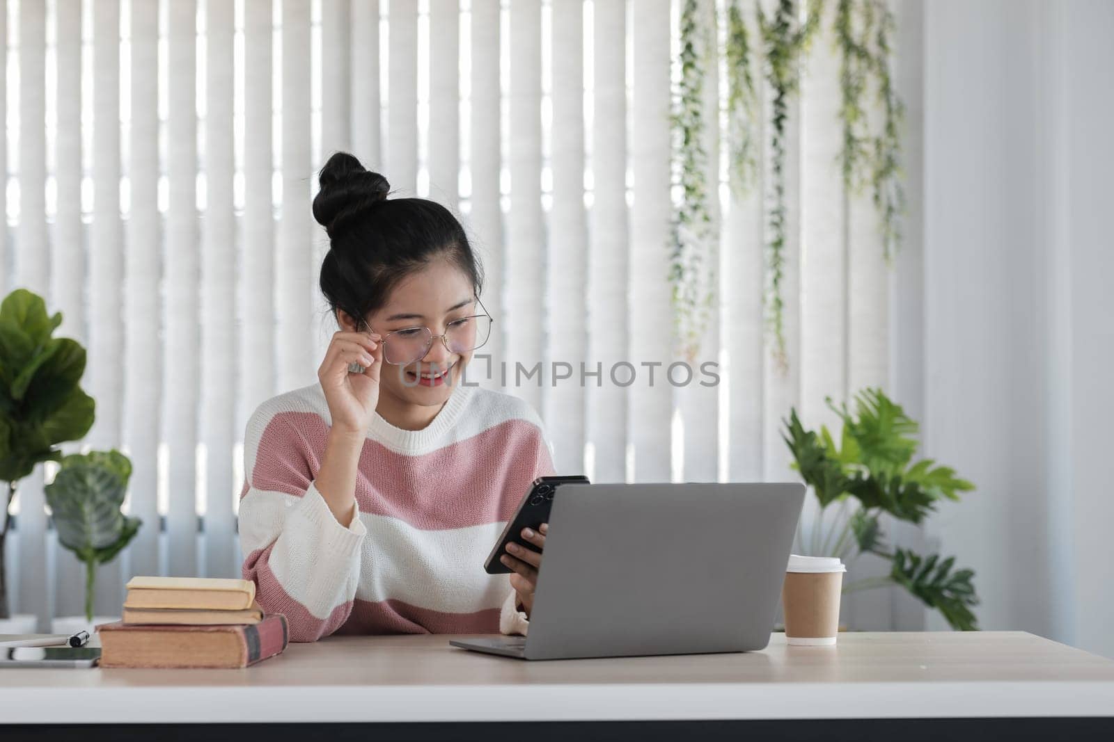 A young woman studies online using a laptop and smartphone in a modern home office, surrounded by plants and books.