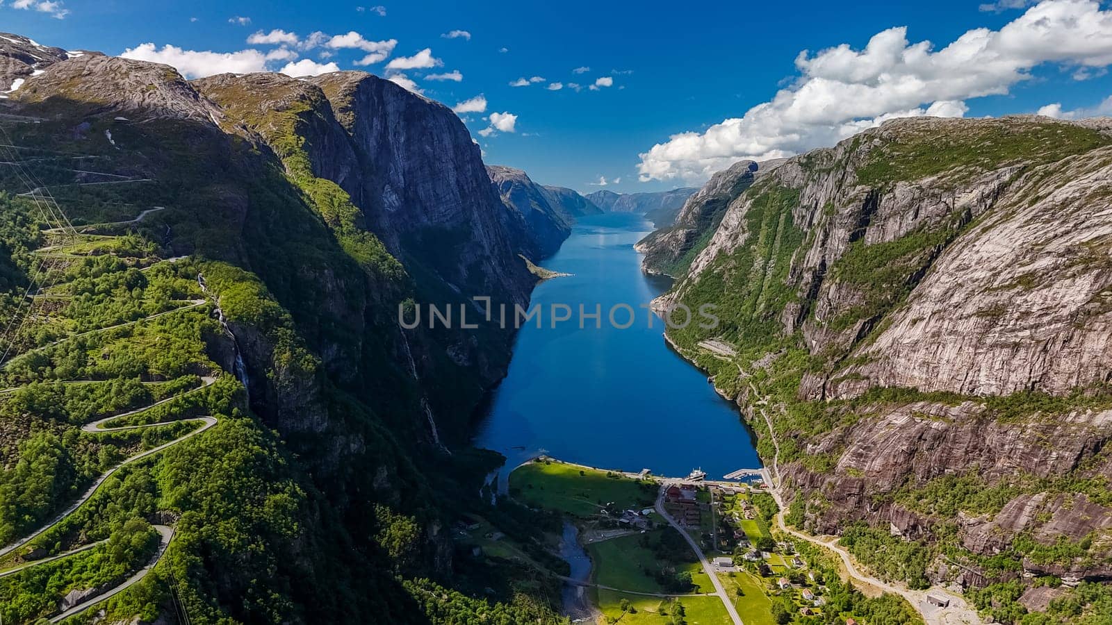 An aerial view of a winding road snaking through the lush green mountains of Norway, with a stunning fjord in the foreground by fokkebok