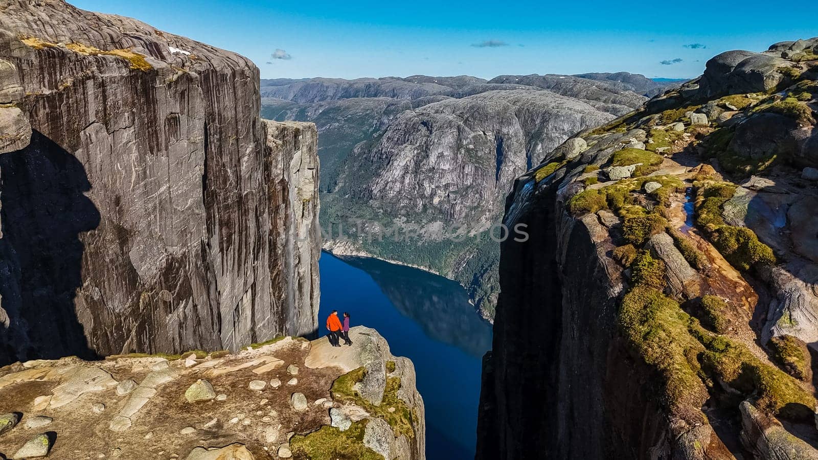 Two individuals stand on the edge of the Kjeragbolten, Norway cliff in Norway, looking out over a breathtaking view of the fjord and surrounding mountains by fokkebok