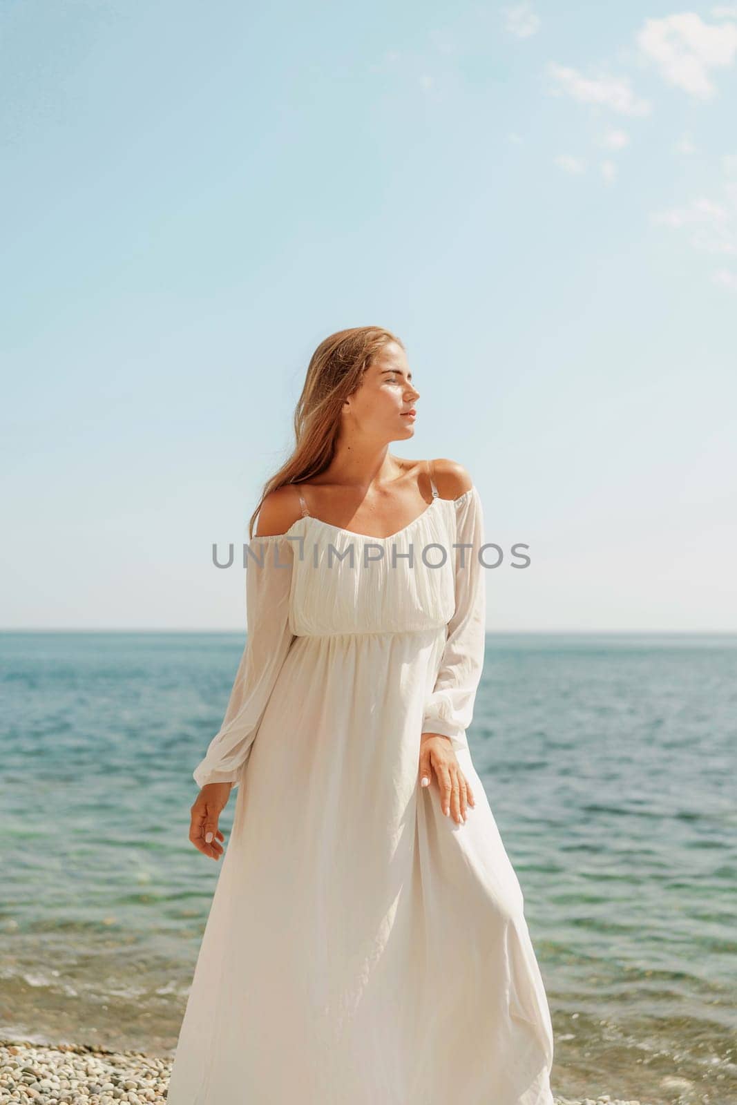 woman white dress stands on a beach, looking out at the ocean. She is in a contemplative or reflective mood, as she raises her hands above her head. Concept of peace and tranquility
