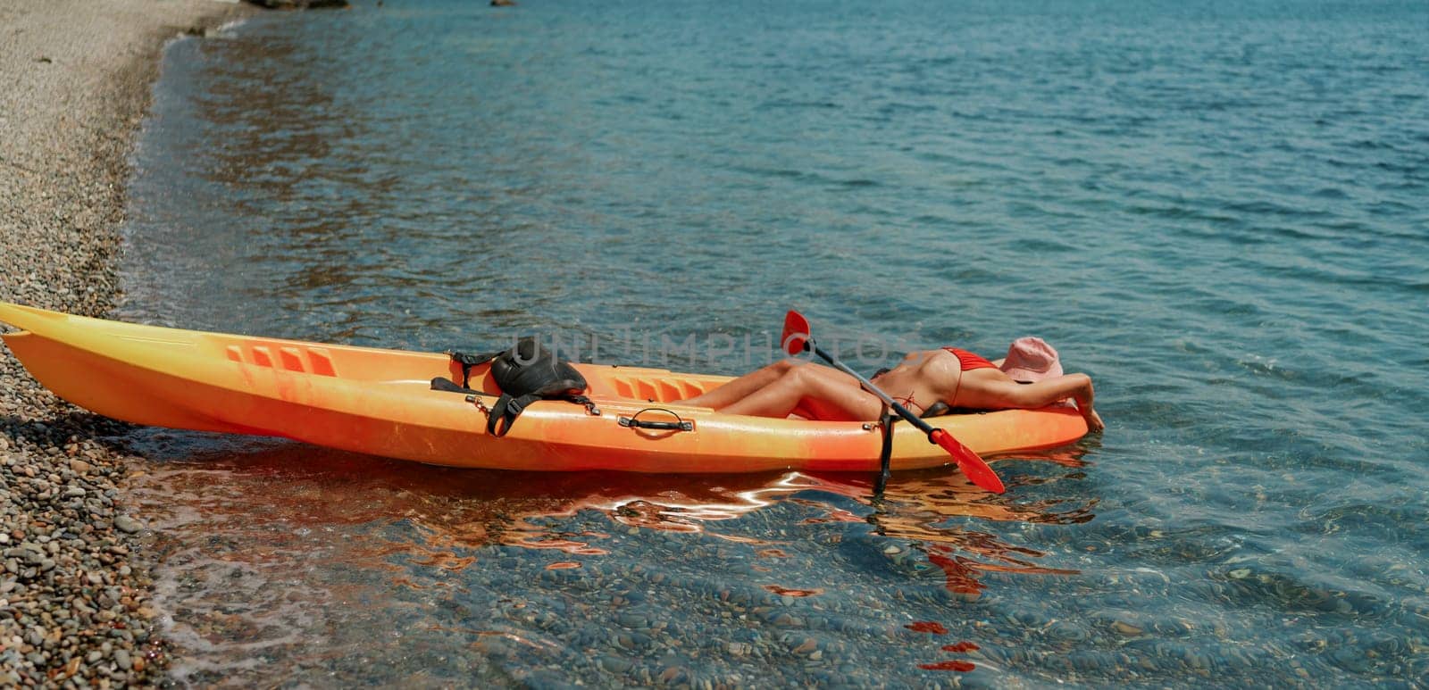 A woman is laying on a kayak in the water. The kayak is orange and has a paddle on it. The woman is wearing a red hat