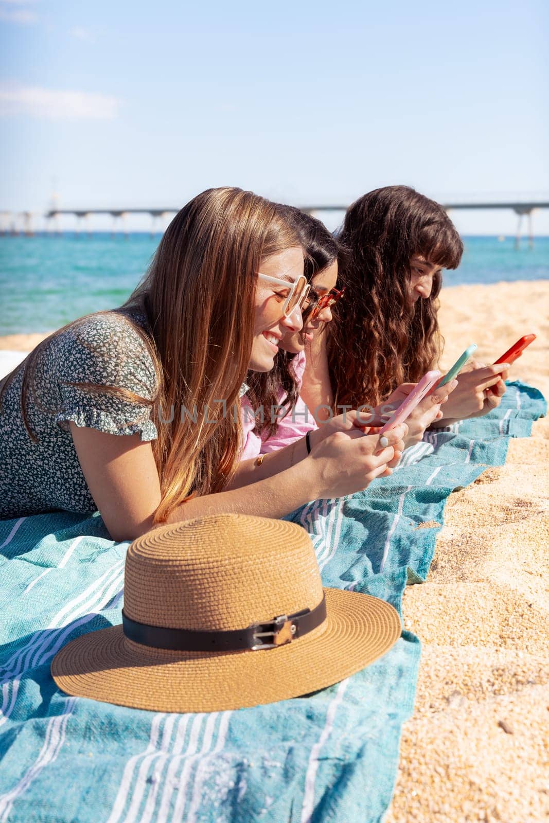 Three smiling friends watching social media while sunbathing on the beach. by mariaphoto3