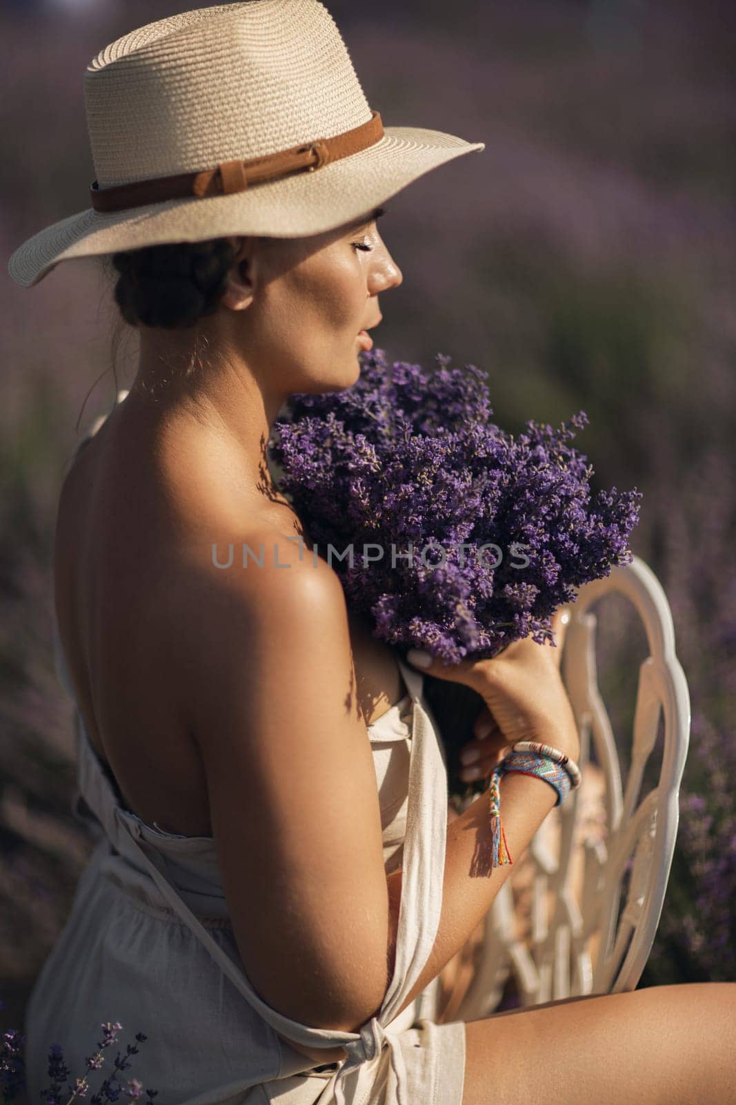 A woman is sitting in a field of lavender flowers and wearing a straw hat. She is smiling and holding a bouquet of flowers. Scene is peaceful and serene, as the woman is surrounded by the beauty of nature.