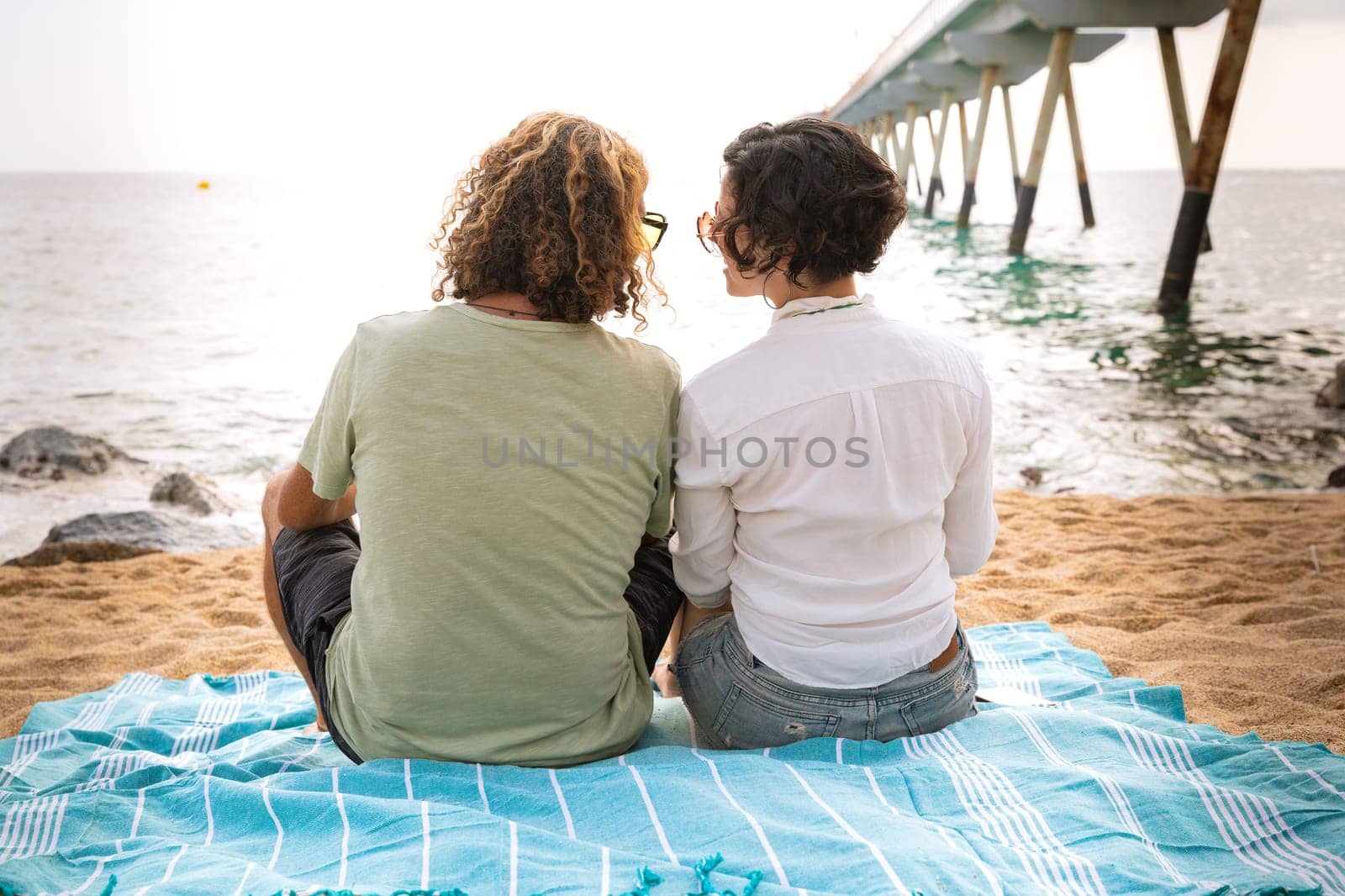 Rear view two happy friends sitting on the beach on vacation,looking at the sea. by molesjuny