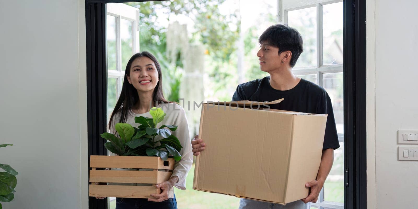 A joyful couple carrying boxes and plants as they move into their new house, symbolizing new beginnings and a fresh start in a modern home.