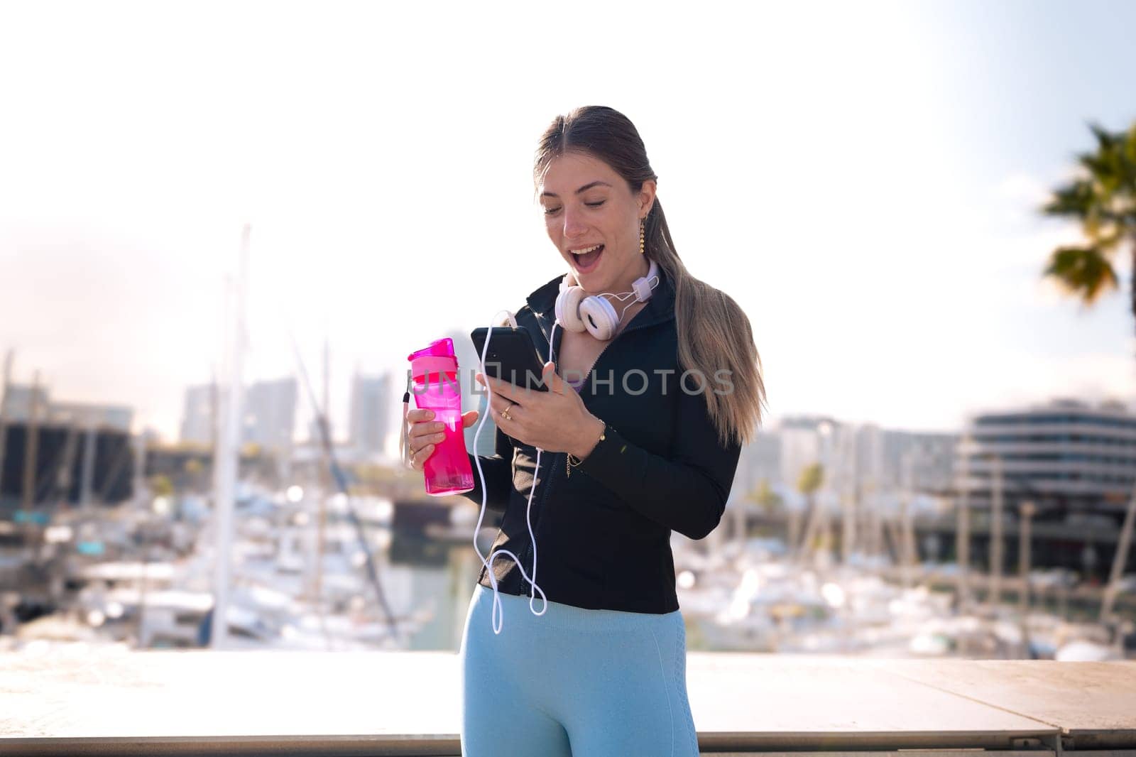 Cheerful girl using an app on a cell phone and holding a sports bottle.