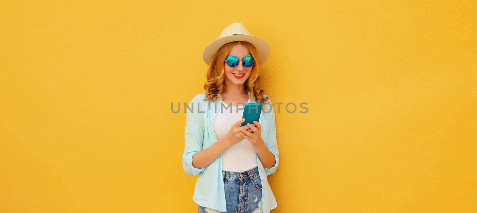 Summer vacation, happy traveler young woman with phone, modern tourist girl looking at smartphone in straw hat on yellow studio background