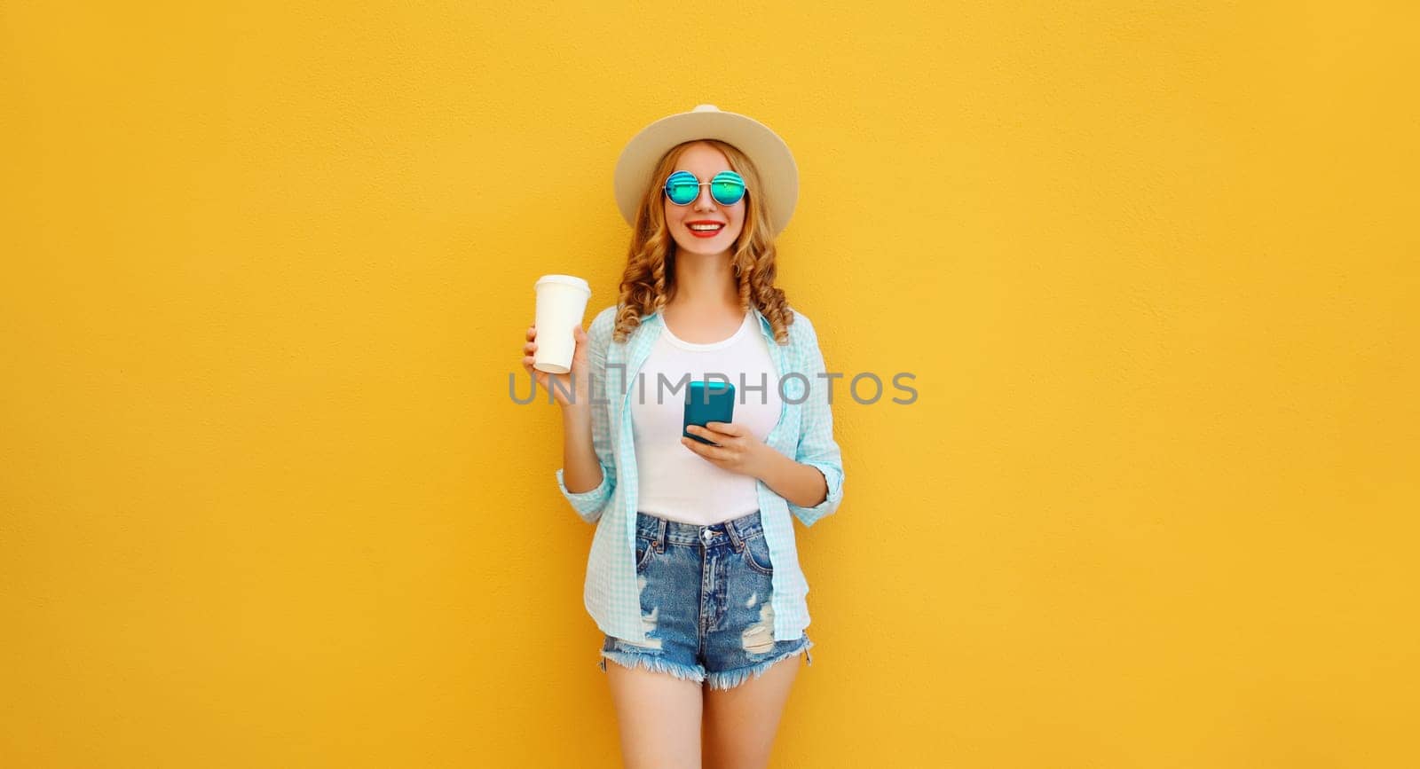 Summer stylish happy young woman with phone, cool girl using smartphone holding cup of coffee dressed tourist straw hat standing on yellow background