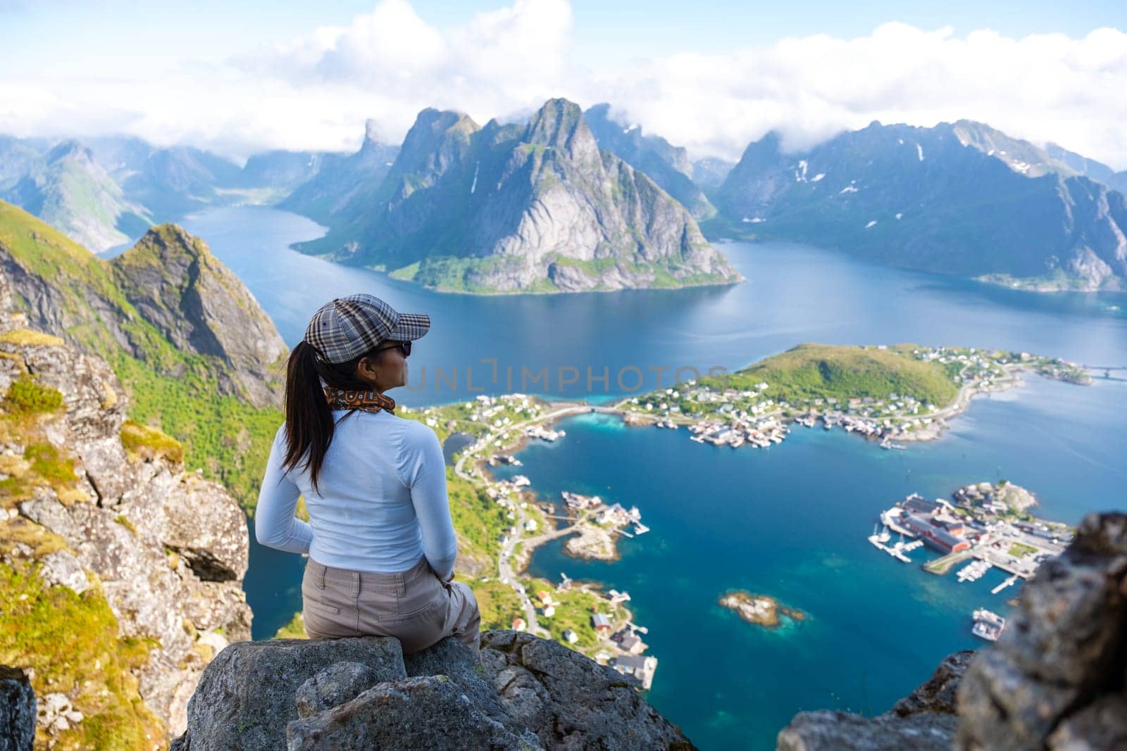 A woman sits on a rocky cliff overlooking a stunning fjord landscape in Norway, enjoying the breathtaking views of the mountains and the sparkling blue waters. Reinebringen, Lofoten, Norway