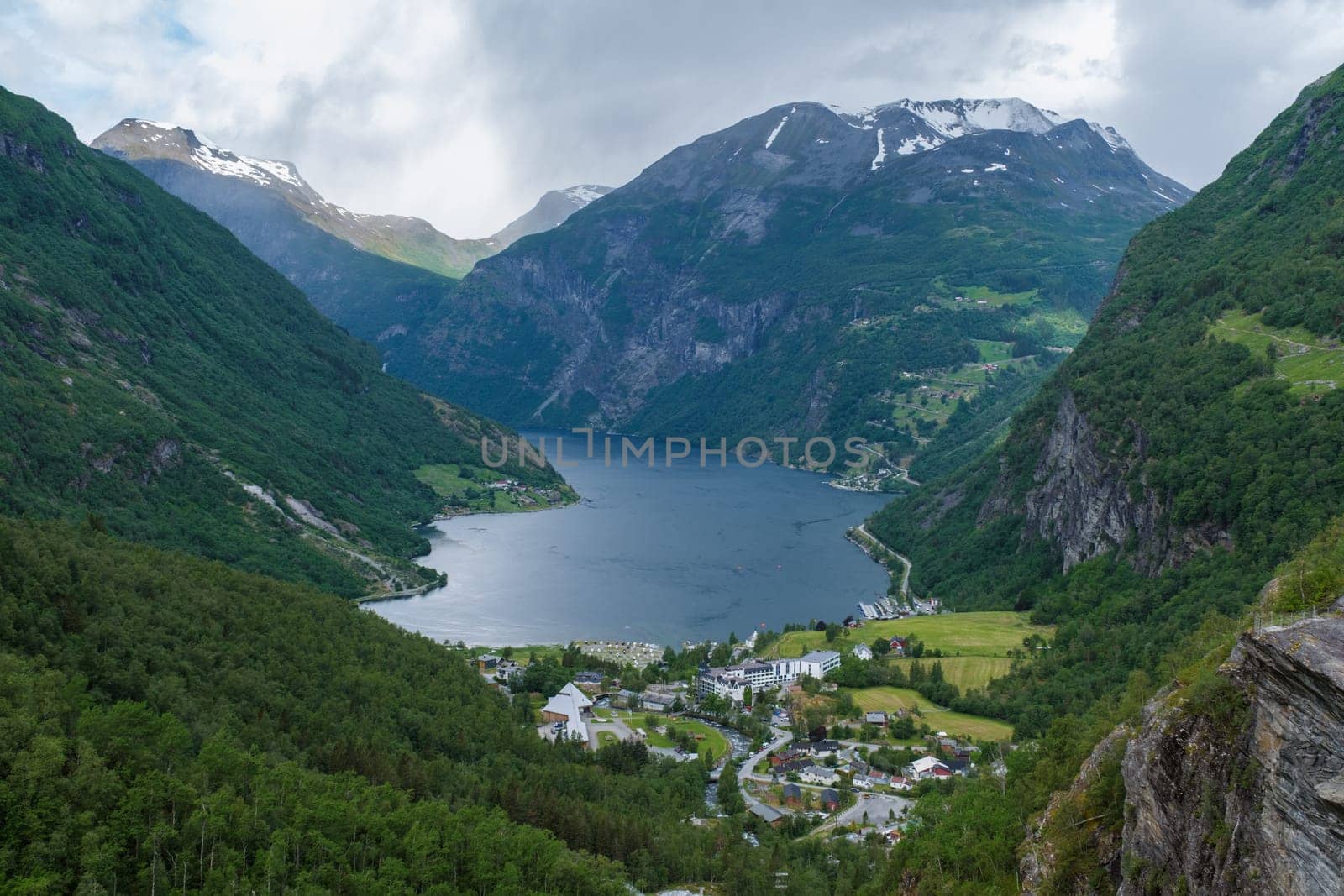 A panoramic view of a picturesque Norwegian fjord, showcasing towering mountains, lush greenery, a winding river, and a small village nestled at the foot of the mountains. Geiranger fjord Norway