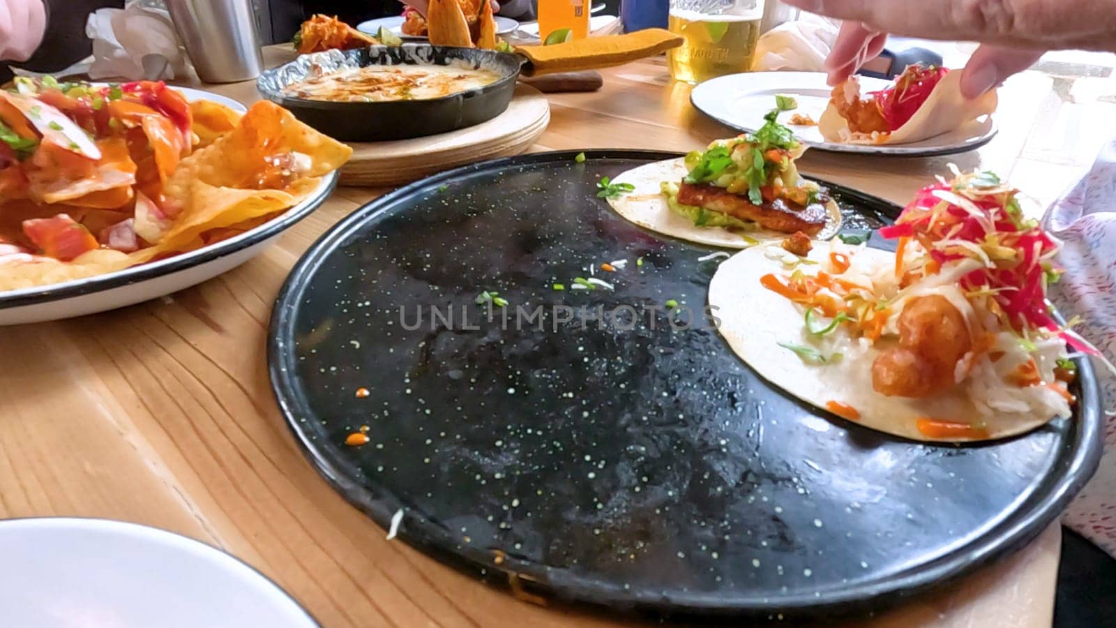 Castle Rock, Colorado, USA-June 12, 2024-Slow motion-Close-up view of a hand picking up a taco from a plate at a restaurant table. The scene features a variety of colorful and appetizing dishes, including nachos topped with cheese and vegetables.