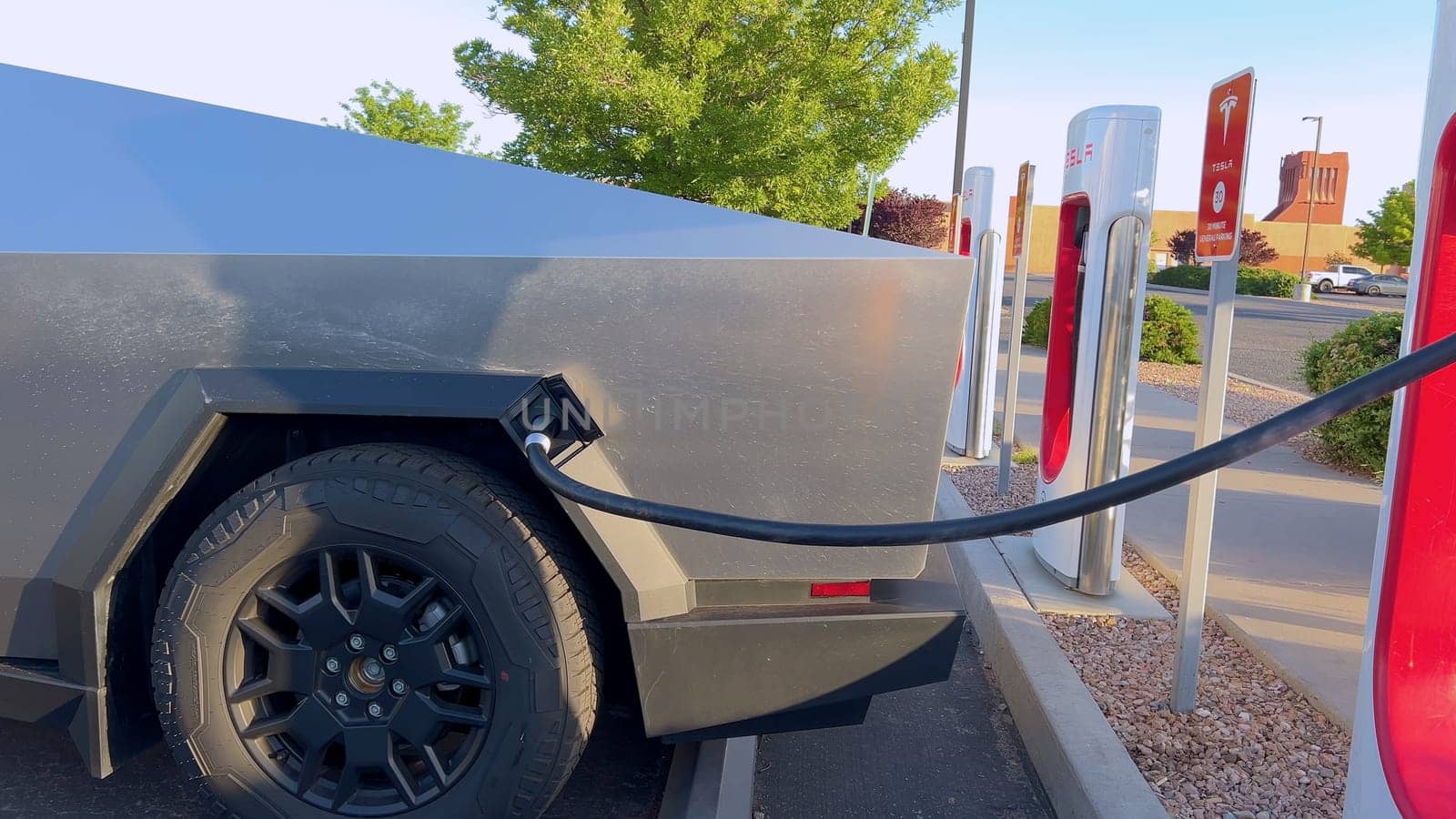 Santa Fe, New Mexico, USA-June 10, 2024-Slow motion-A Tesla Cybertruck is connected to a Tesla Supercharger at an outdoor charging station. The sleek, angular design of the Cybertruck is highlighted by the sunlight, emphasizing its modern and futuristic appearance.