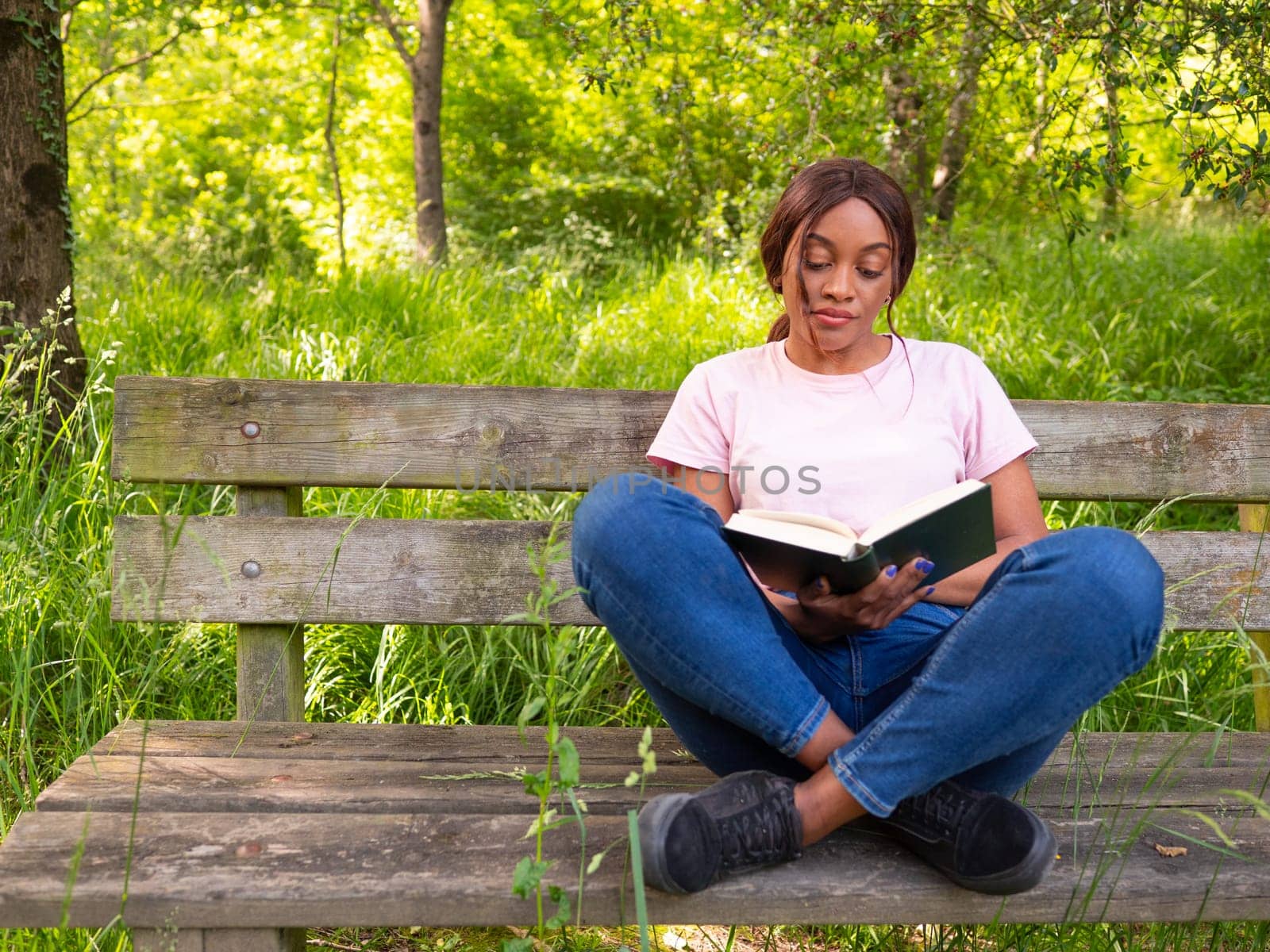 Young black woman sitting on a park bench reading a book. by Ceballos