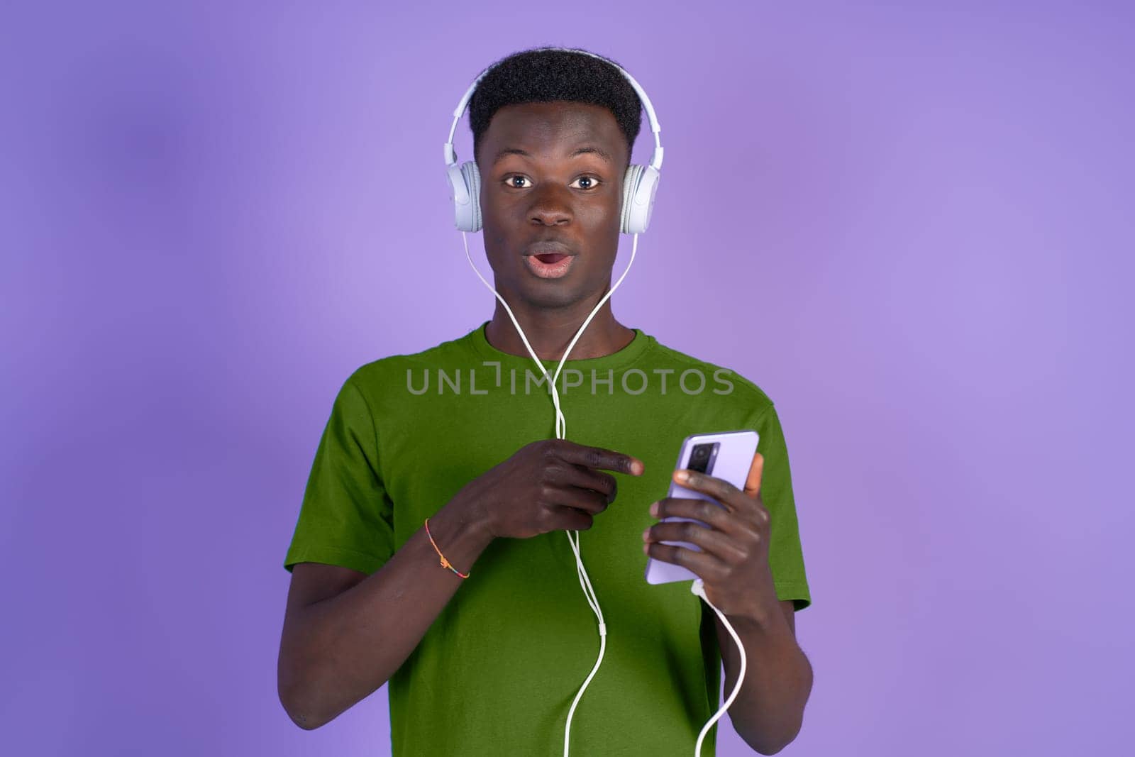 Young black man amazed while listening to music with headphones and a mobile phone on a violet background, with copyspace. by Ceballos