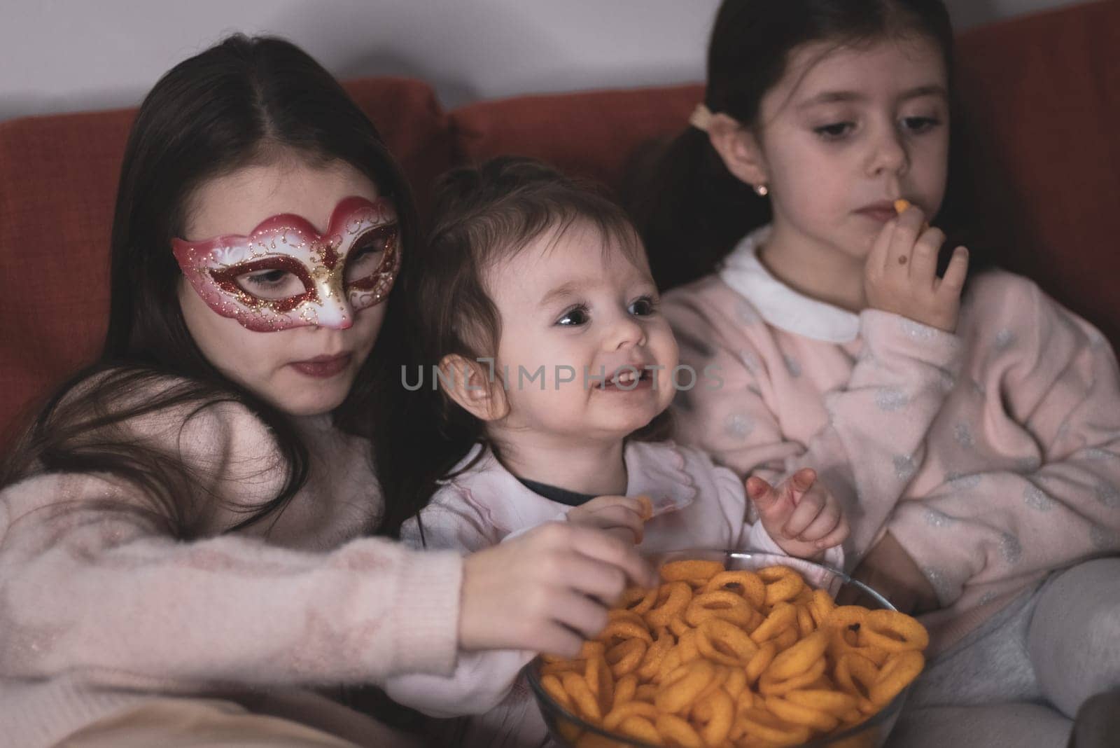 Three beautiful caucasian girls sisters are sitting on the sofa,eating corn rings and emotionally watching a movie in the room in the evening,close-up side view.The concept of family watching movies.