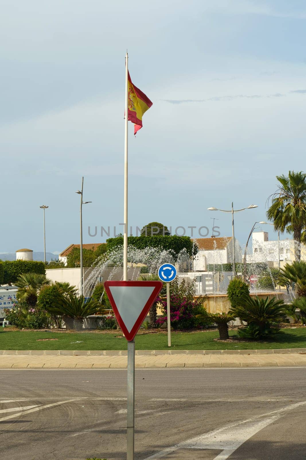 Almeria, Spain - May 25, 2023: A Spanish flag flies high above a roundabout in Almeria, Spain. A yield sign stands in the center of the roundabout, with lush greenery and palm trees surrounding it.