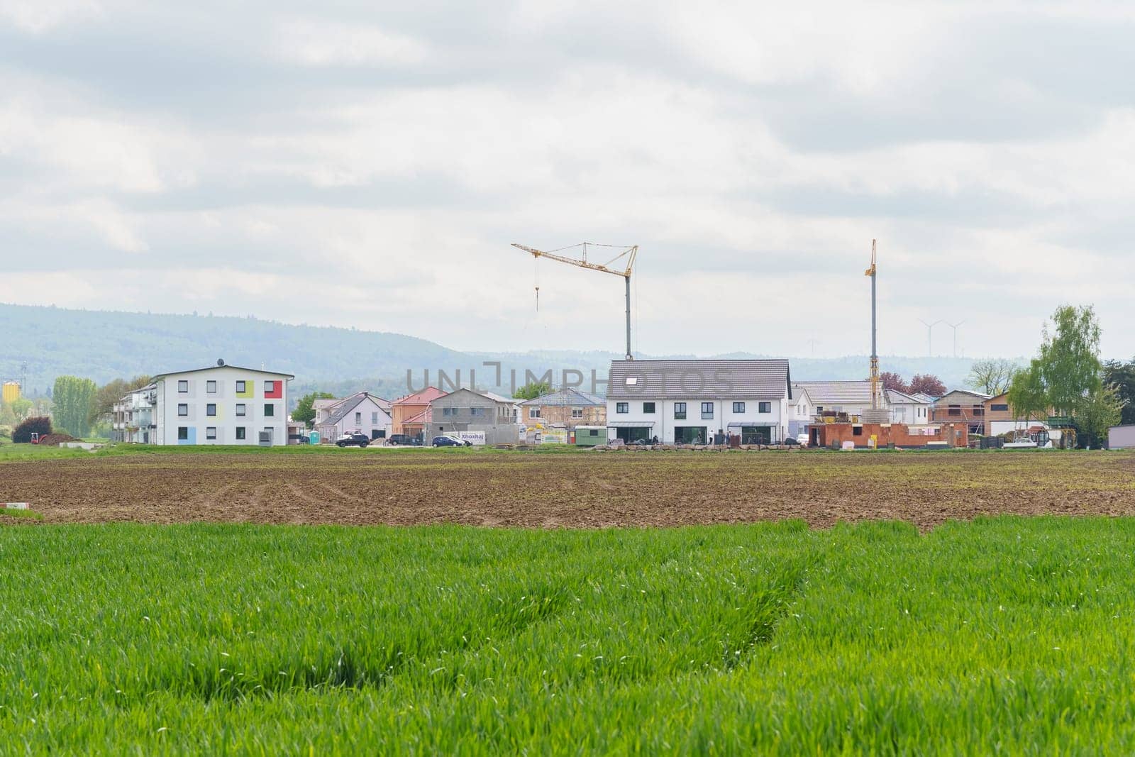 Schuttern, Germany - April 29, 2023: A view of a new housing development with a construction crane in the background, a field of grass in the foreground, and a cloudy sky above.
