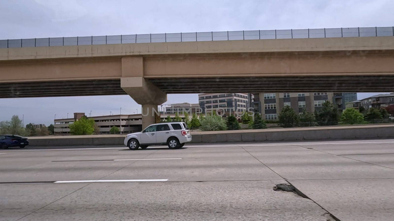 Denver, Colorado, USA-June 11, 2024-Slow motion-A scene from I-25 Highway in Denver, Colorado, capturing vehicles in motion, including a white SUV and a silver sedan. The backdrop features office buildings and lush green trees under a cloudy sky, illustrating typical highway traffic in this bustling city.