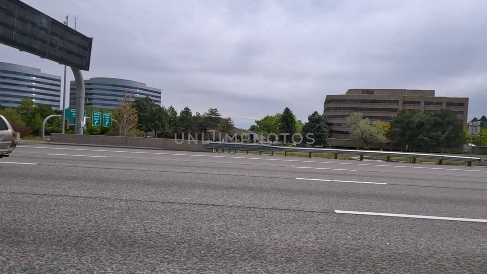 Denver, Colorado, USA-June 11, 2024-Slow motion-A scene from I-25 Highway in Denver, Colorado, capturing vehicles in motion, including a white SUV and a silver sedan. The backdrop features office buildings and lush green trees under a cloudy sky, illustrating typical highway traffic in this bustling city.