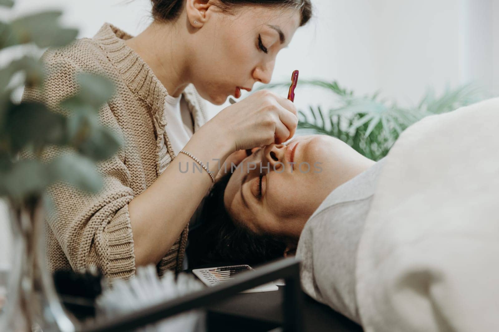 Portrait of one young beautiful Caucasian brunette girl cosmetologist who glues artificial eyelashes using two tweezers on the left eye of a female client lying on a cosmetology table in a home office on a spring day, close-up side view. Eyelash extension concept, beauty industry.