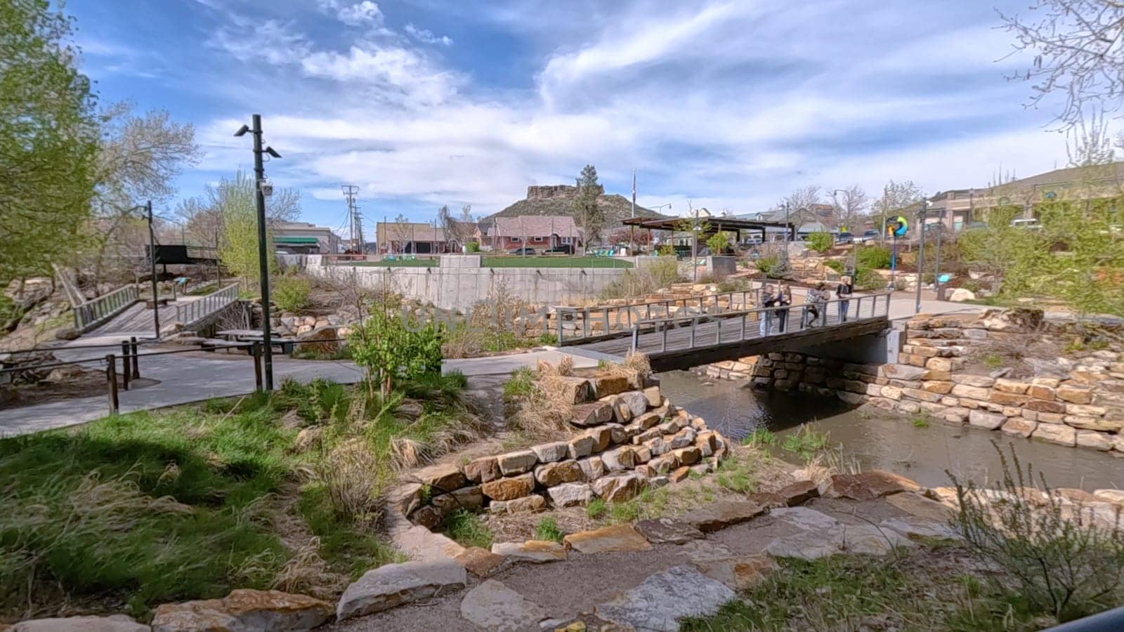 Castle Rock, Colorado, USA-June 12, 2024-Slow motion-A picturesque view of Downtown Castle Rock, Colorado, featuring a serene river flowing through the urban landscape. The scene includes a charming bridge, lush greenery, and modern buildings in the background under a bright blue sky with scattered clouds. The blend of nature and urban development highlights the unique character of this vibrant community.