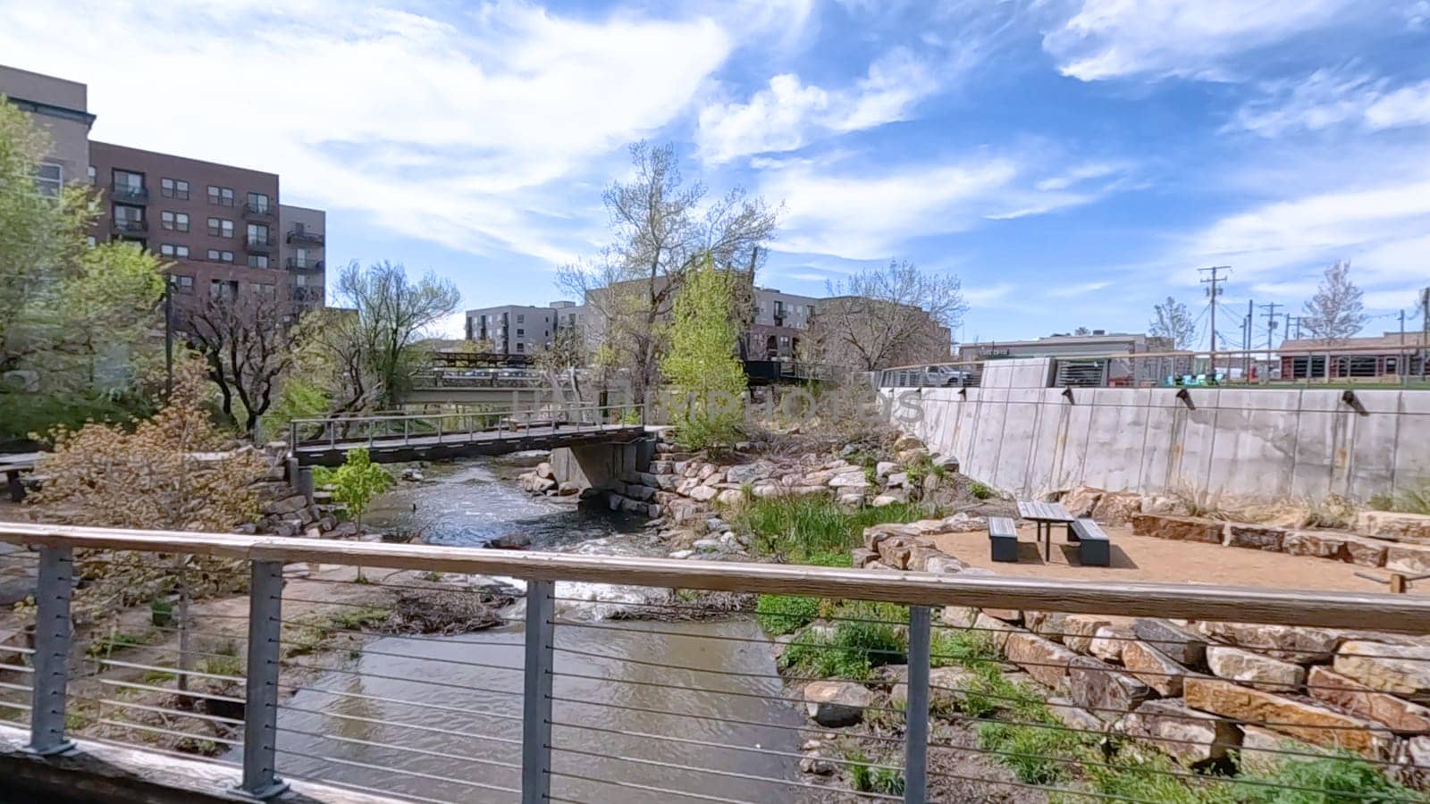 Castle Rock, Colorado, USA-June 12, 2024-Slow motion-A picturesque view of Downtown Castle Rock, Colorado, featuring a serene river flowing through the urban landscape. The scene includes a charming bridge, lush greenery, and modern buildings in the background under a bright blue sky with scattered clouds. The blend of nature and urban development highlights the unique character of this vibrant community.