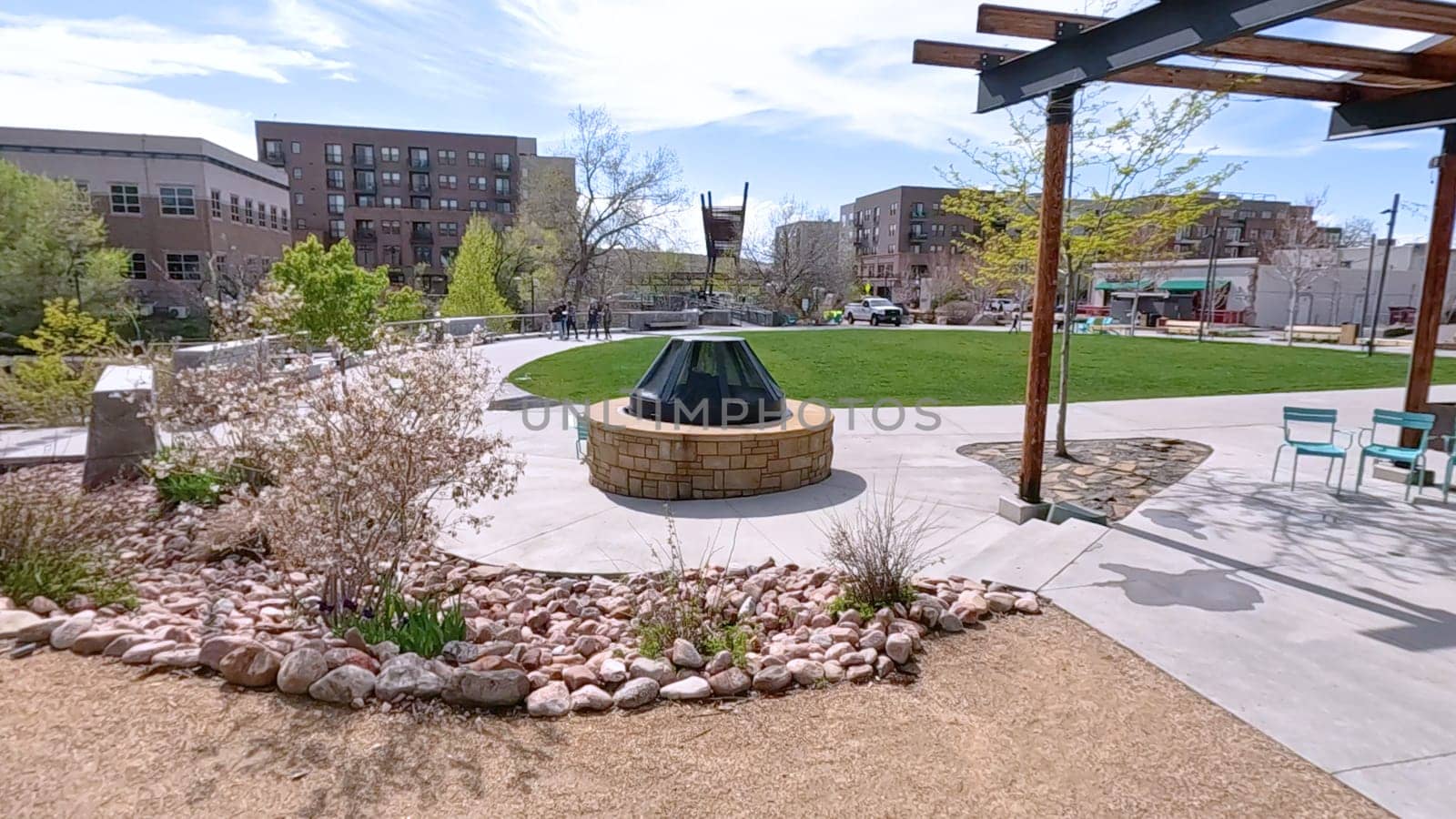 Castle Rock, Colorado, USA-June 12, 2024-Slow motion-A beautiful urban park located in Downtown Castle Rock, Colorado, showcasing modern seating areas, a cozy fire pit, and a lush green lawn. The park features a mix of natural and architectural elements, including a wooden pergola and well-designed pathways, creating an inviting outdoor space for relaxation and community gatherings. Surrounding buildings and vibrant greenery enhance the park charm under a clear blue sky.