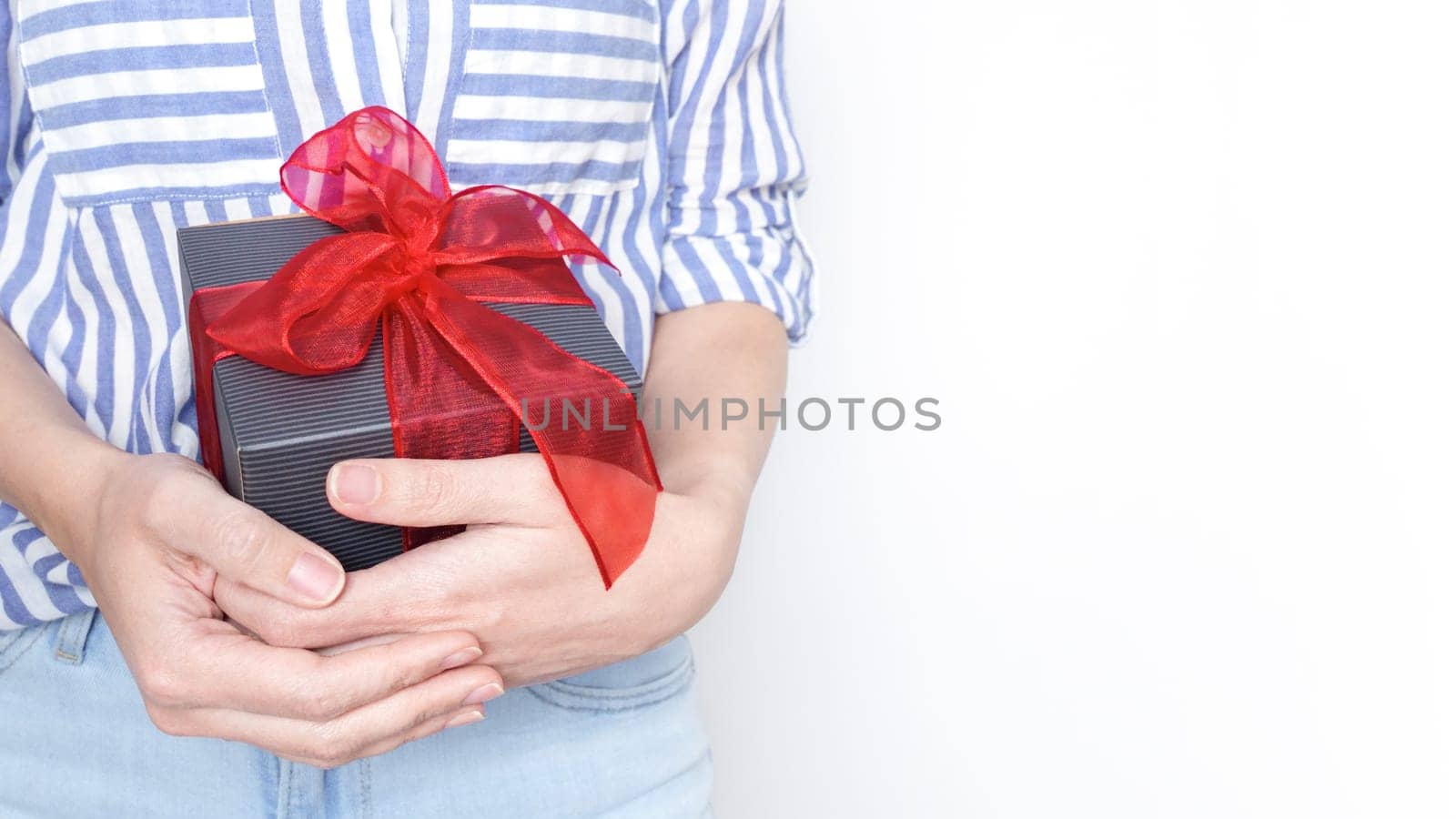 A young woman smiling while holding a beautifully wrapped gift box with a red ribbon. She is wearing a striped shirt and jeans, looking happy. The background is plain white.