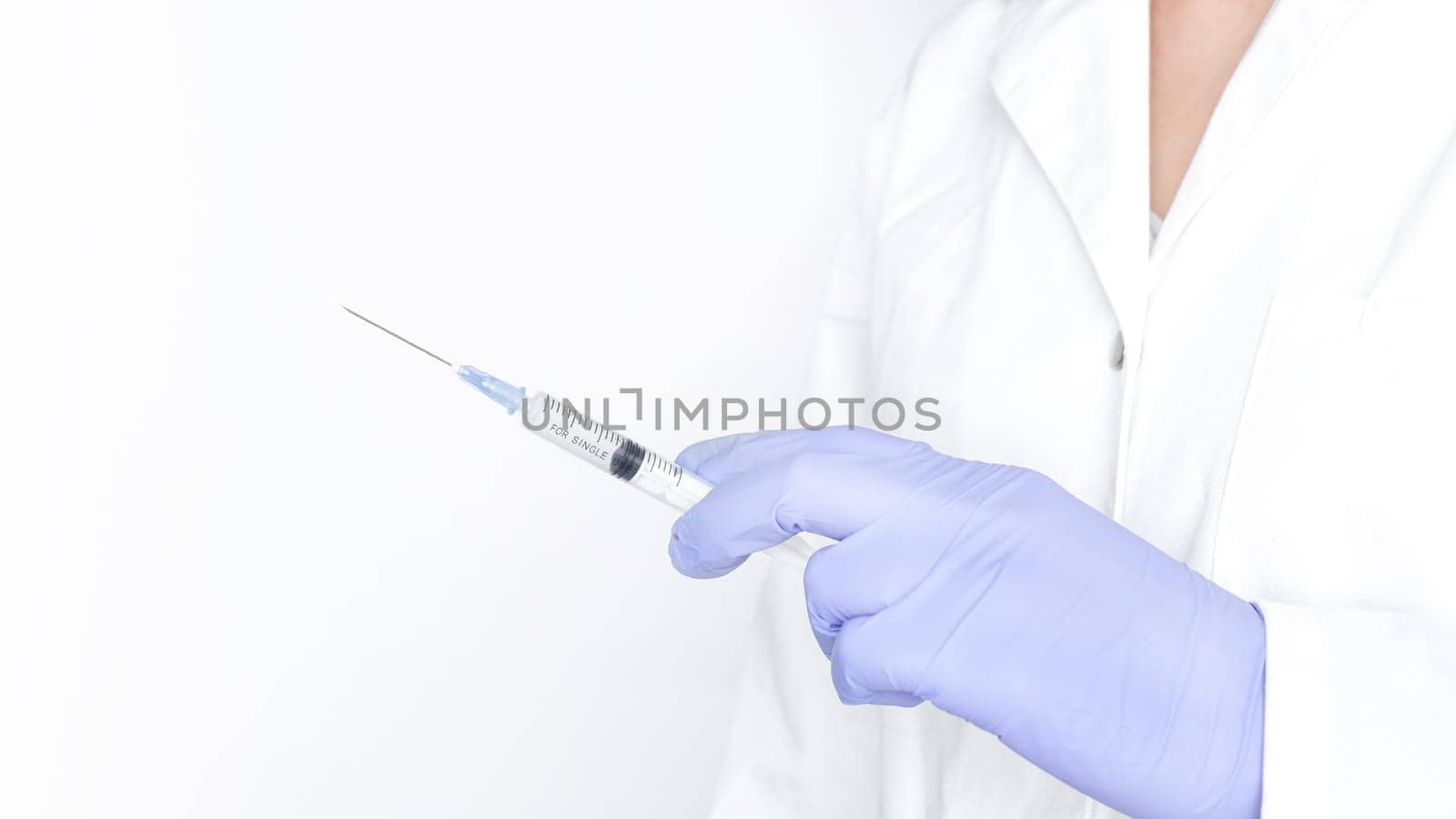 A gloved hand is holding a syringe with a needle. The syringe is filled with a clear liquid. The hand is holding the syringe in a vertical position. The needle is pointing up.