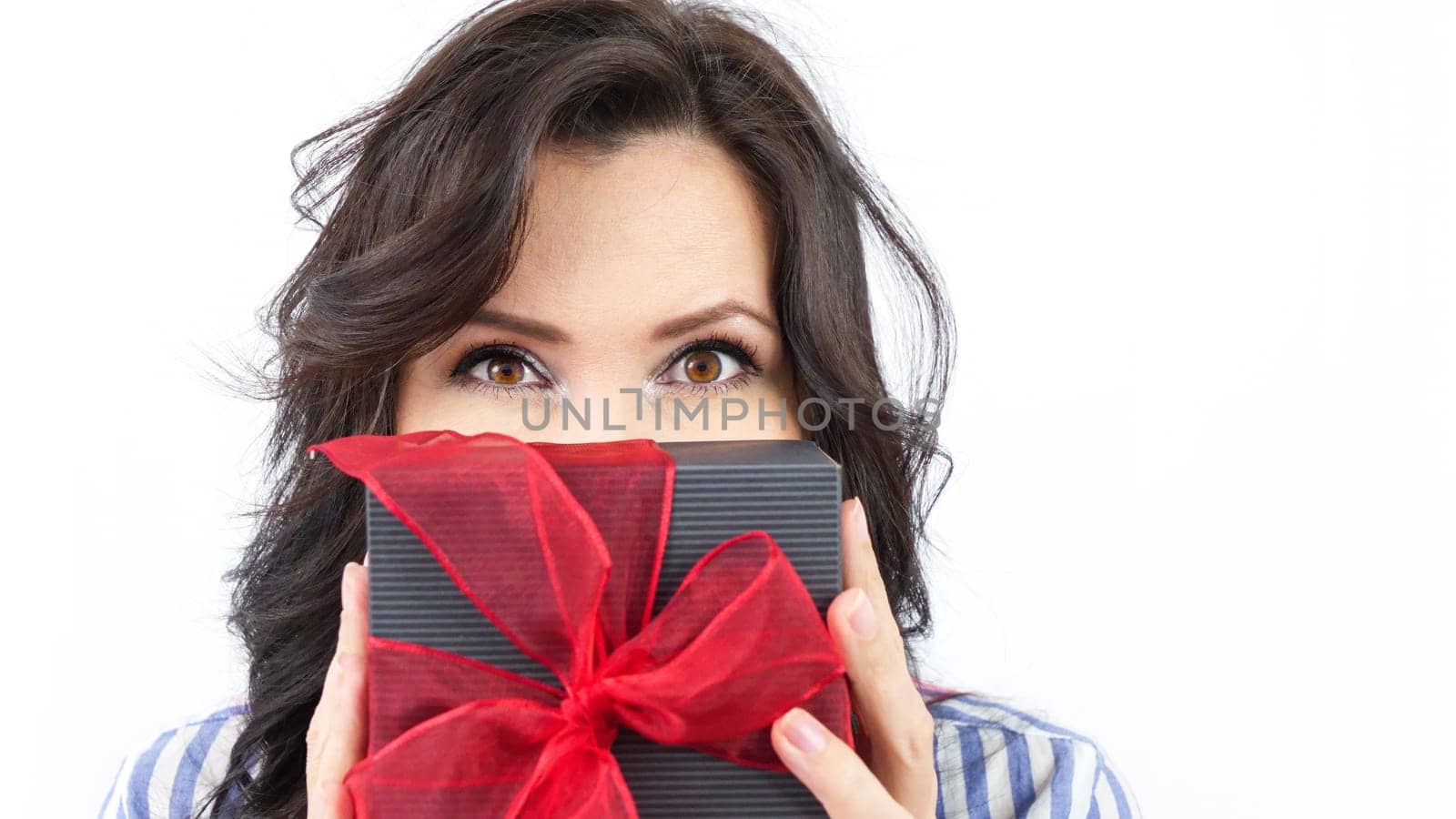 A young woman with long brown hair is holding a black gift box with a red ribbon. She is looking at the camera with a surprised expression. The woman is wearing a blue and white striped shirt.