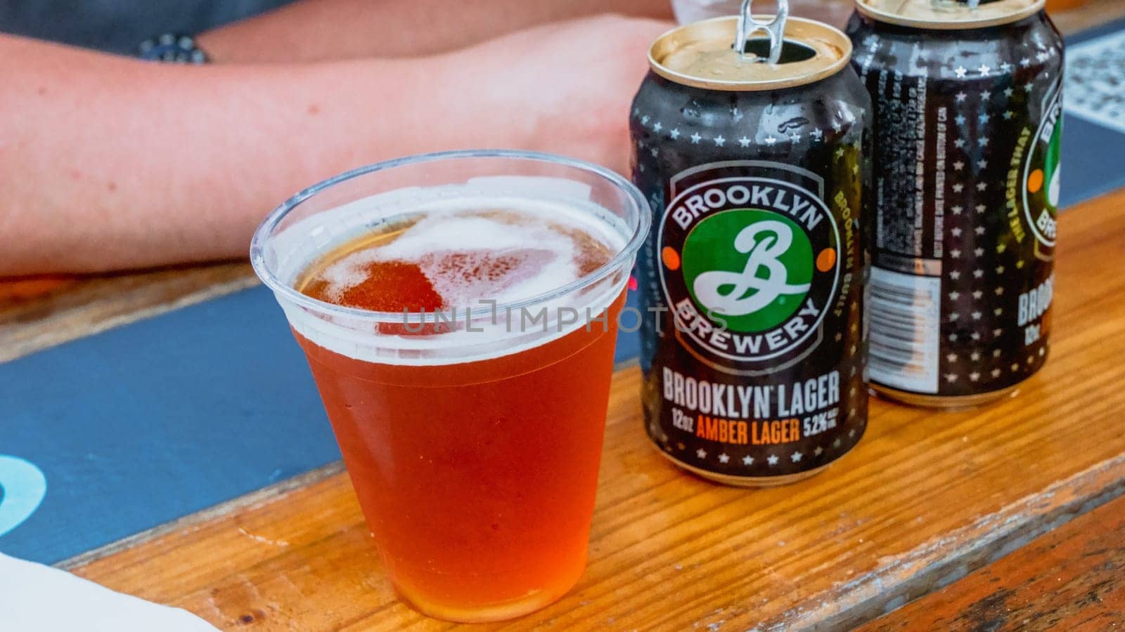 A half-full plastic cup of amber lager sits on a wooden table next to two unopened cans of the same beer. The Brooklyn Brewery logo is visible on the can. Isolated on a wooden background.