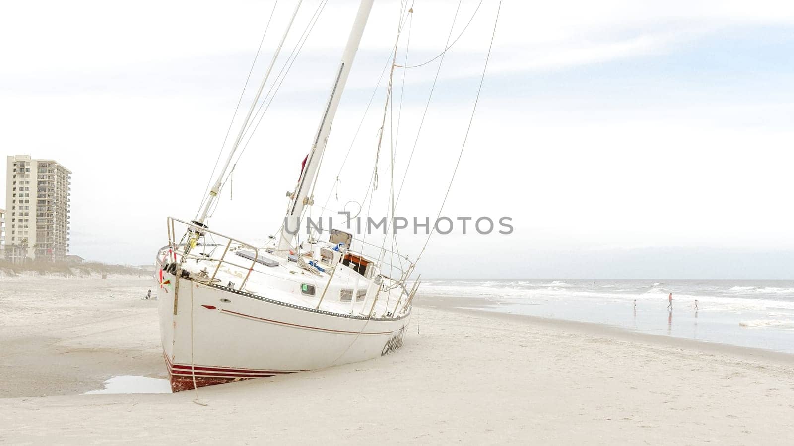 A beached yacht rests on the sandy shore after a hurricane. The damaged vessel sits askew under a gloomy sky as waves crash against the coastline, creating a scene of destruction and despair.