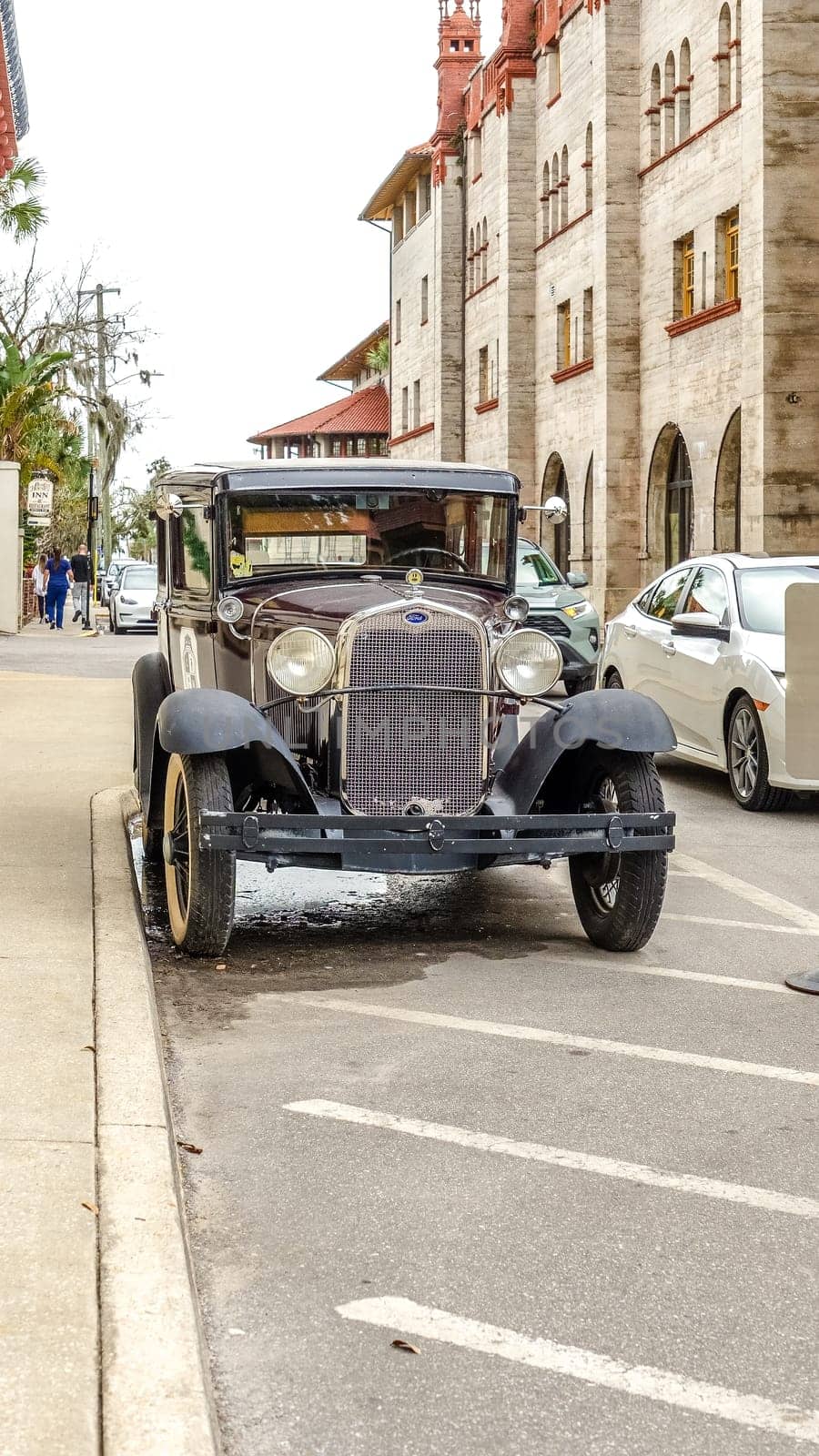 A vintage black car parked on a town road with two pedestrians walking by. The car features shiny black paint, chrome bumpers, and white wall tires, set against buildings and palm trees.