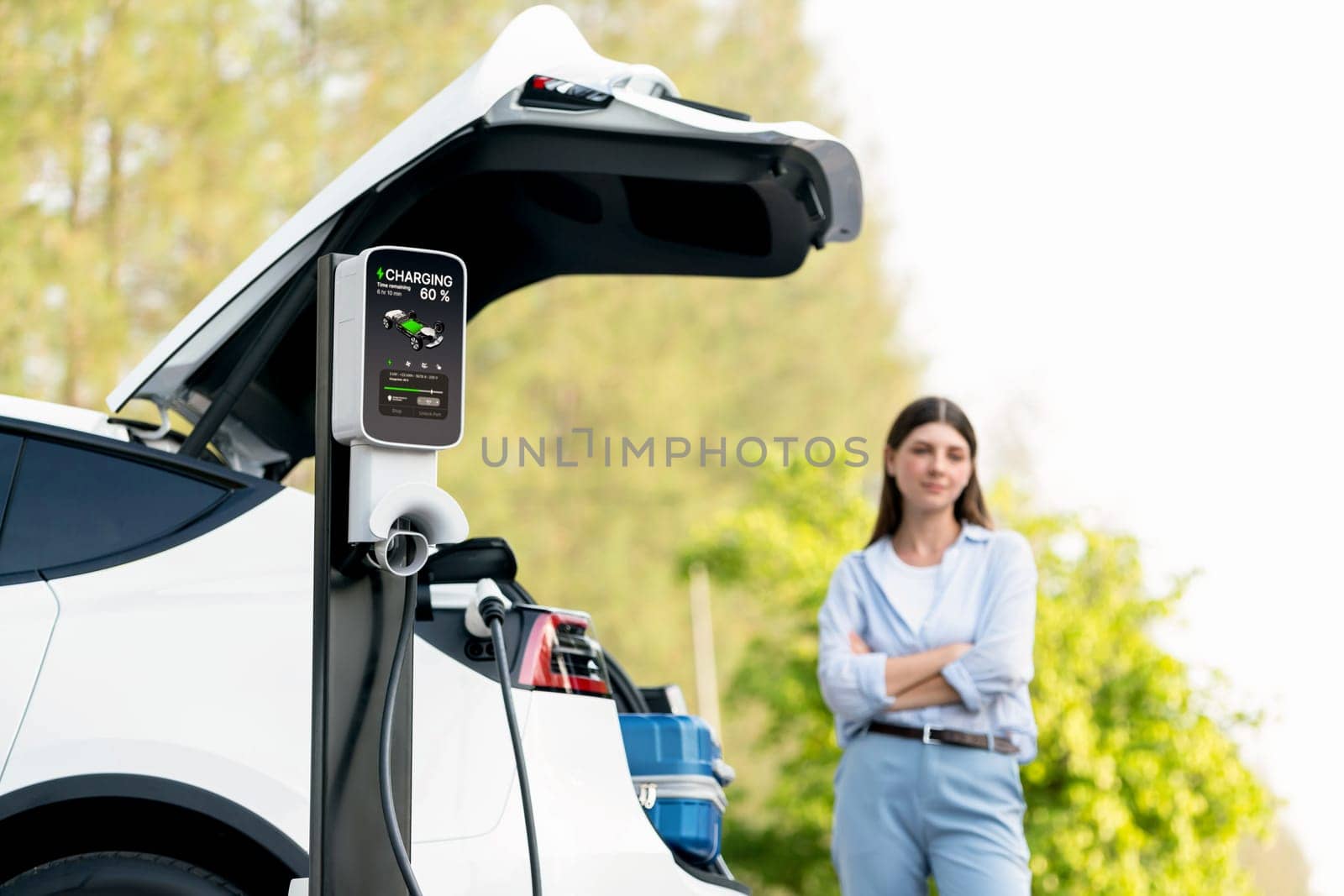 Young woman recharging battery for electric car during autumnal road trip travel EV car in autumnal forest. Eco friendly travel on vacation during autumn with electric vehicle. Exalt