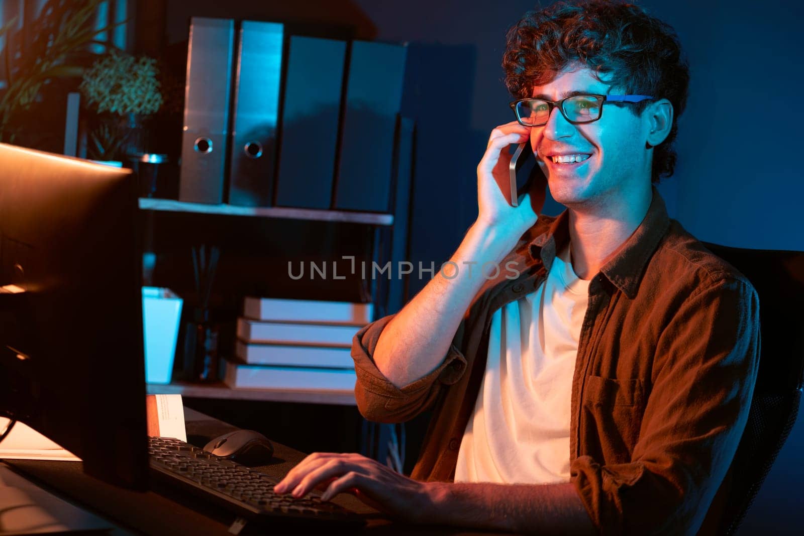 Young businessman calling on smartphone to customer or coworker, looking at computer screen of creative presentation surround work desk at neon modern office. Concept of new next project. Gusher.