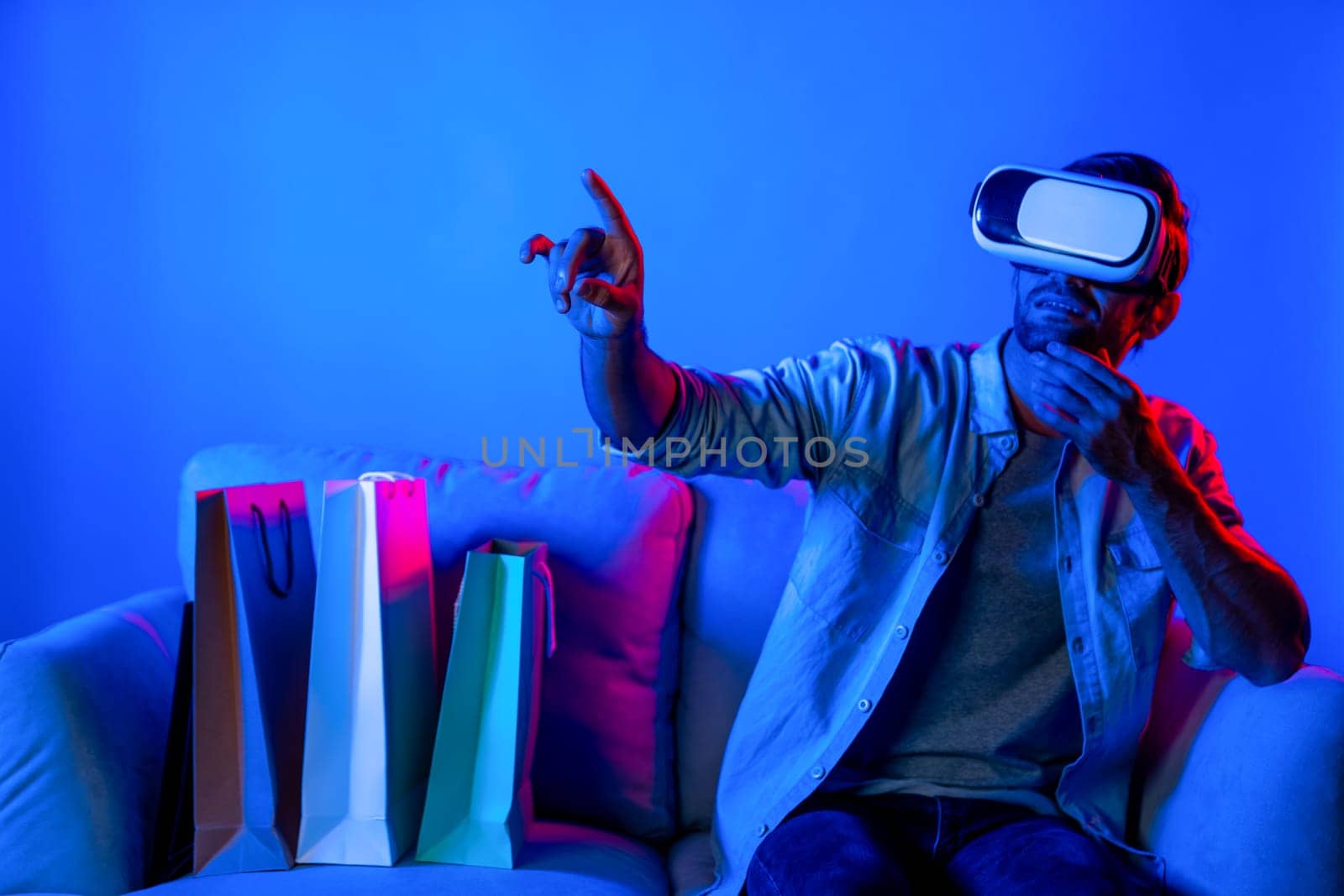 Caucasian man with vr glass posing pointing finger while sitting at sofa with shopping bags. Person selecting and doing shopping online by using VR technology and virtual goggle at home. Deviation.