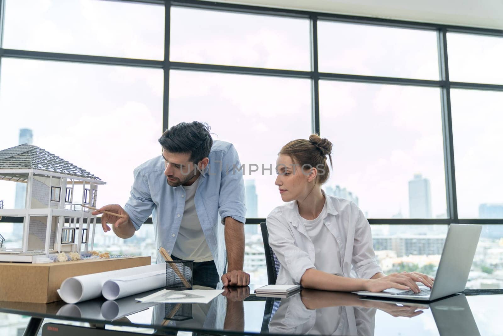 Young skilled project manager working by using laptop while caucasian engineer inspect house mode, using project plan at modern office or architect studio. Civil engineering, creative design. Tracery.