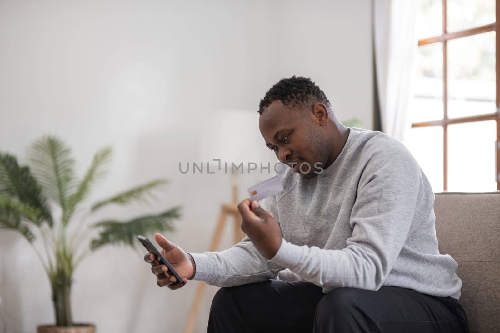 African American man sitting on a sofa at home, looking at a credit card while holding a smartphone, bright window light