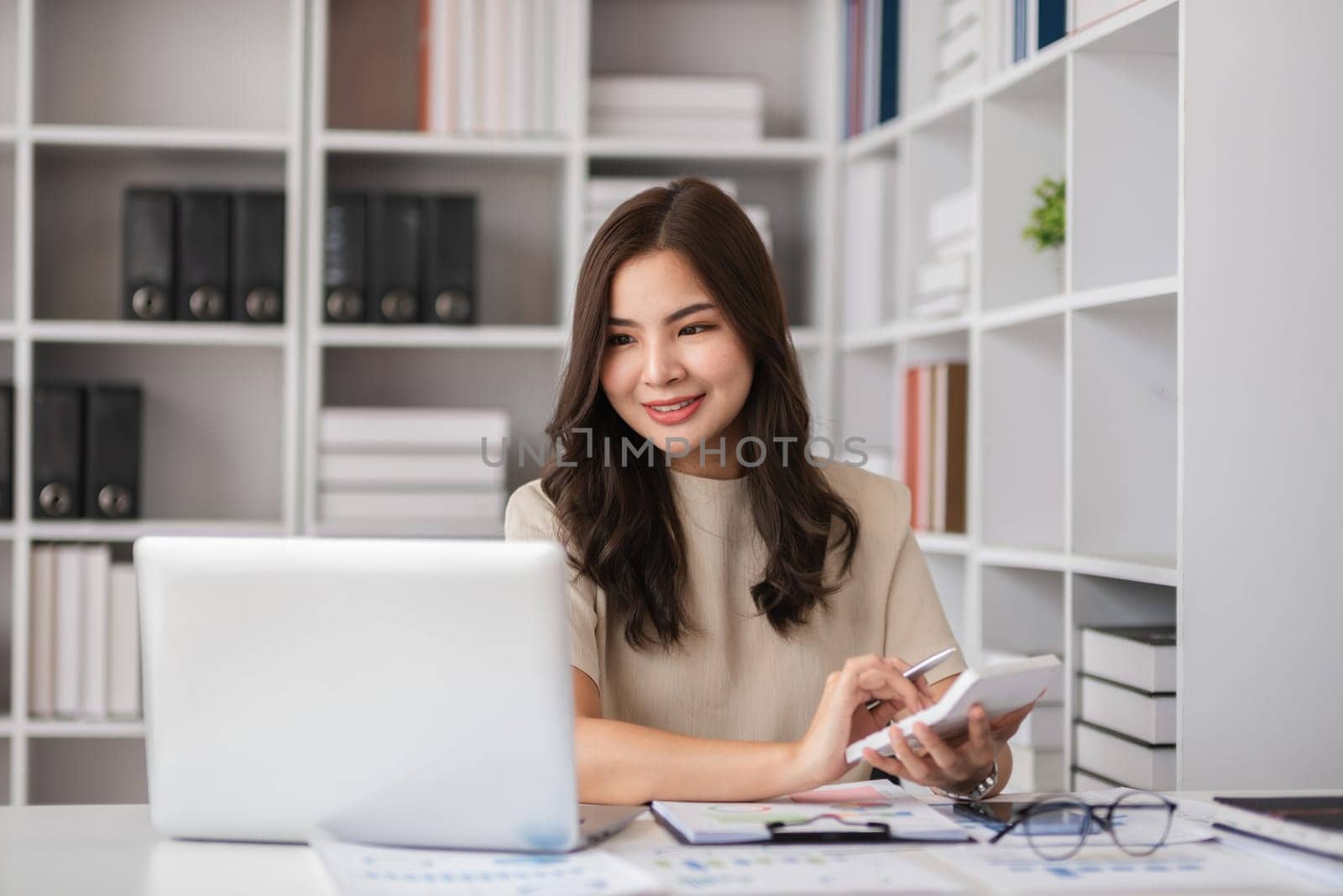 Young Female Accountant Working from Home with Laptop and Calculator in Modern Home Office Setting by wichayada