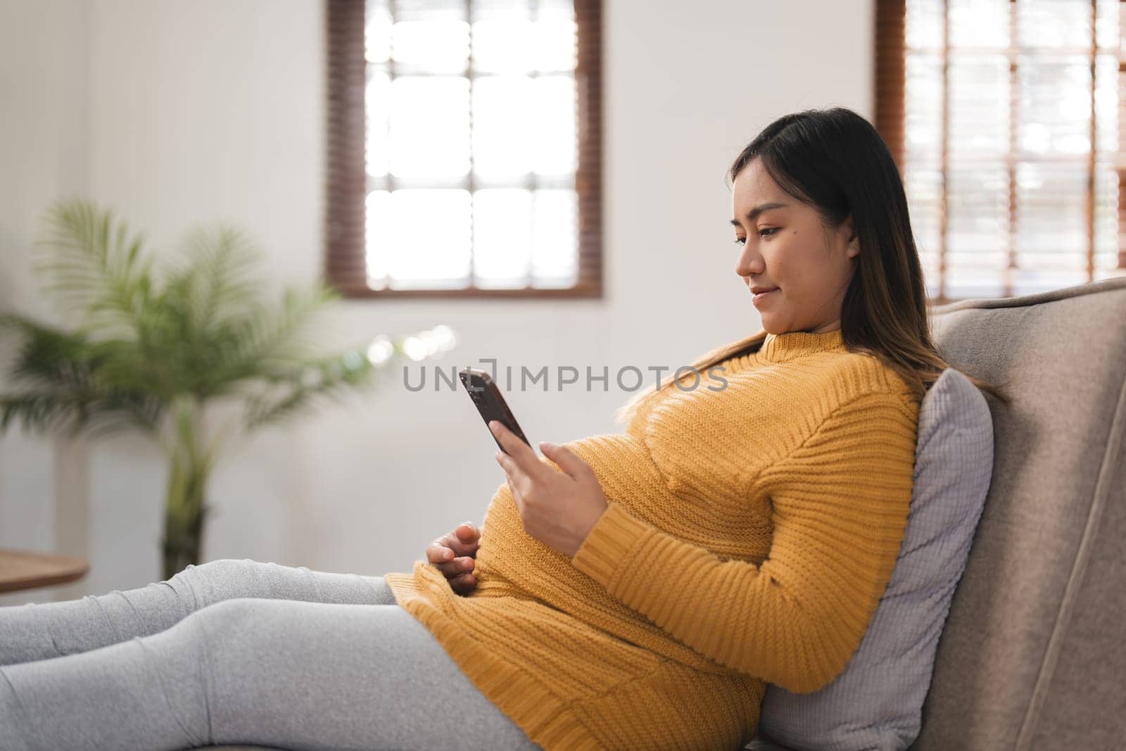 Pregnant woman sitting on a sofa in a modern living room, using a smartphone, wearing comfortable maternity clothing, with natural light and a cozy home environment.