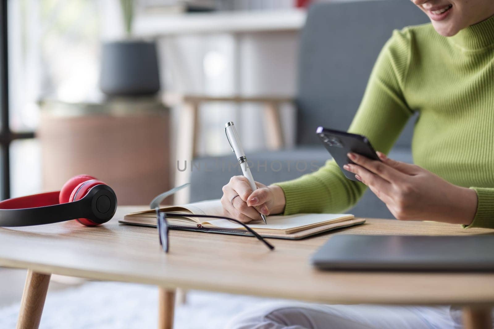 Close-up of Asian woman multitasking with smartphone, notebook, and headphones in modern office, concept of efficiency by wichayada