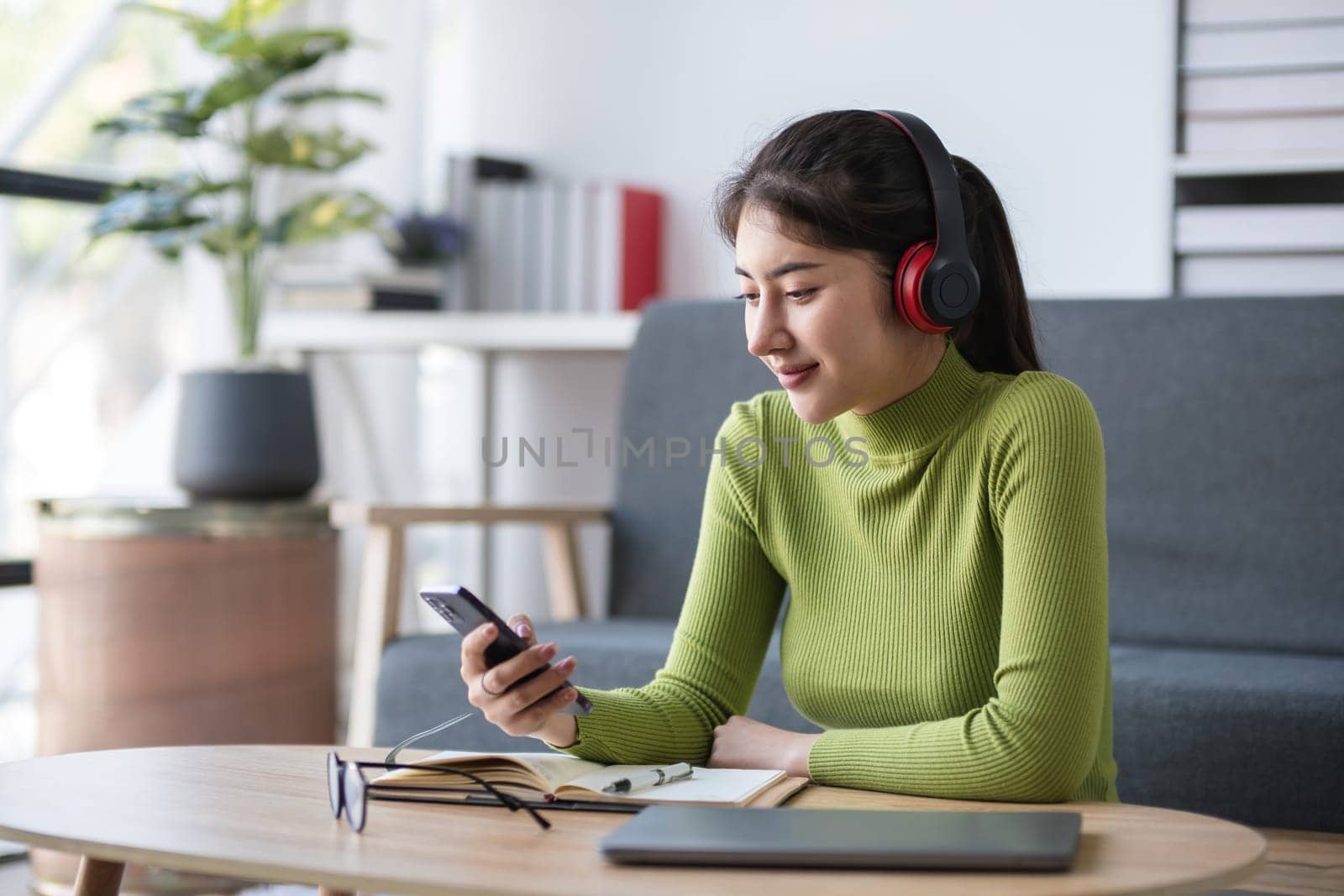 Asian woman checking smartphone while working with headphones in modern office, concept of connectivity.