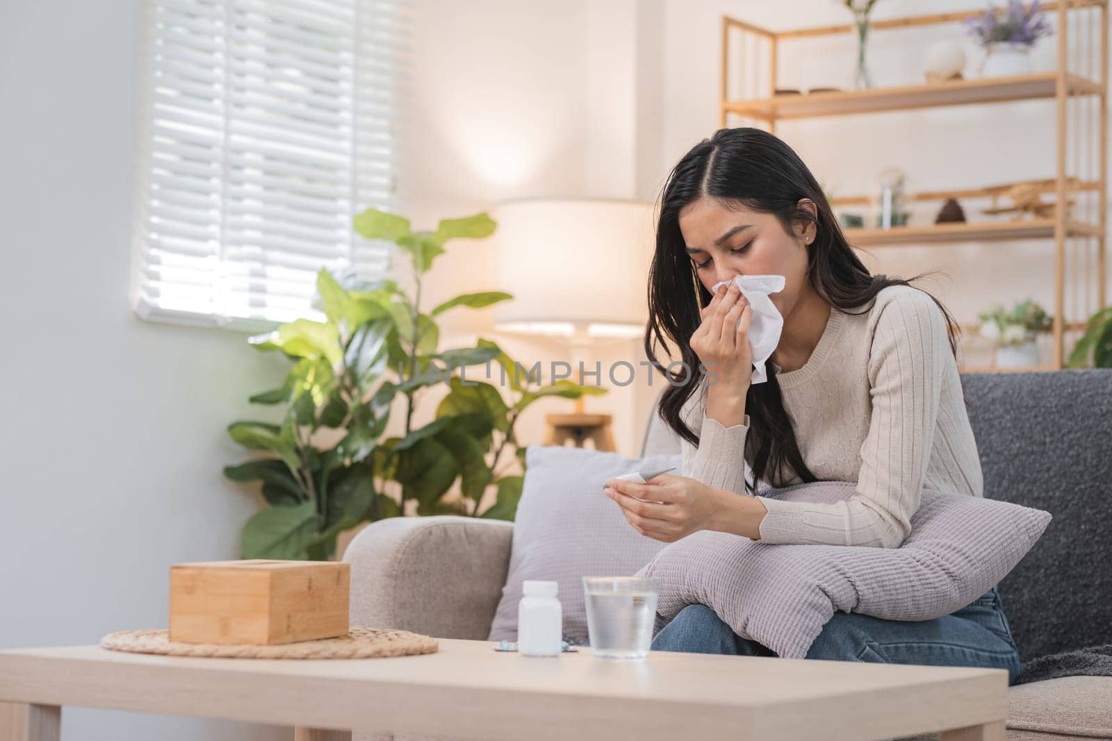 Young Woman Sitting on Couch Blowing Nose with Tissue in Bright Living Room with Plants and Shelves in Background by wichayada