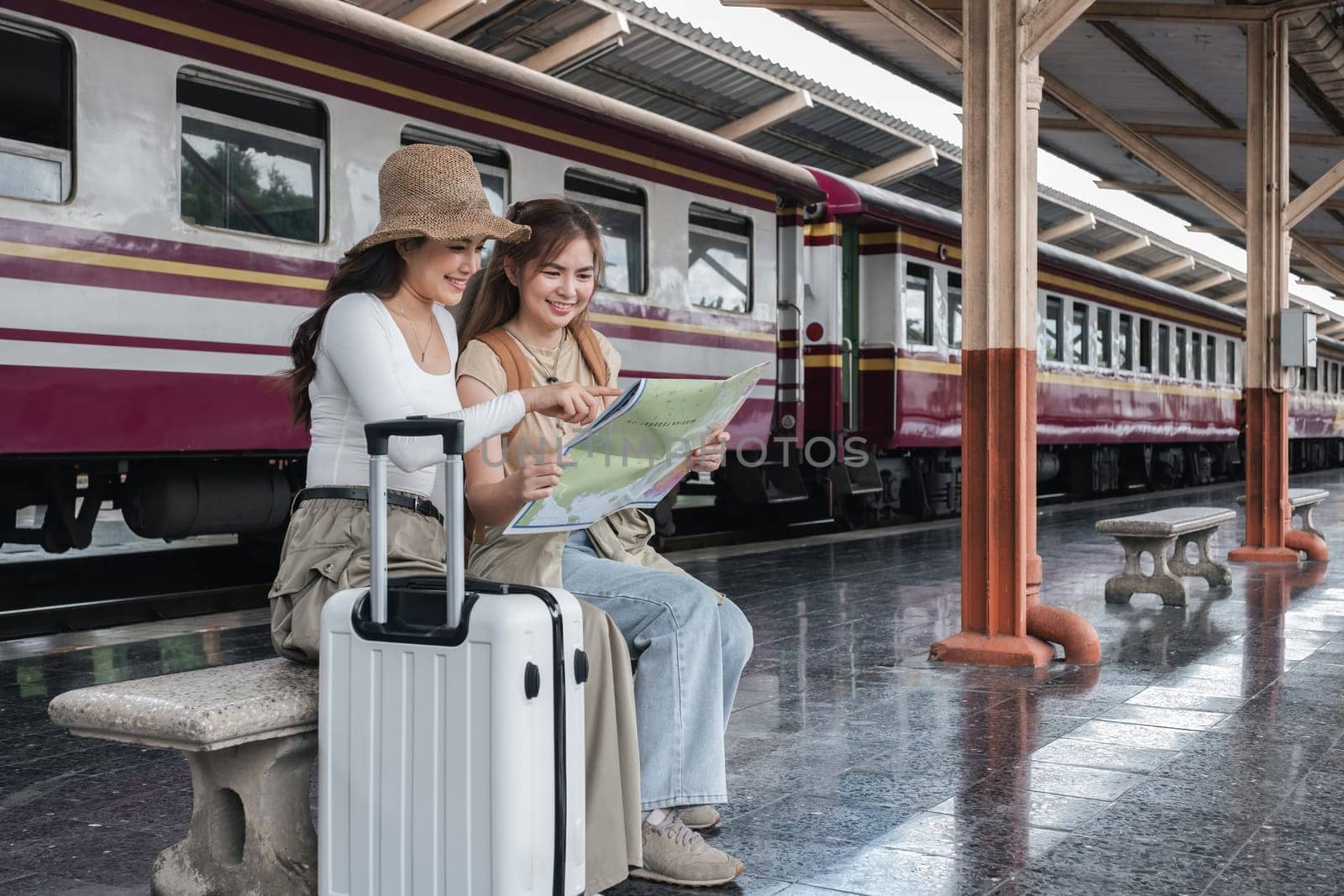 Two young Asian women sitting on a bench at a train station, holding a map and pointing, with a suitcase beside them, planning their travel route with excitement.